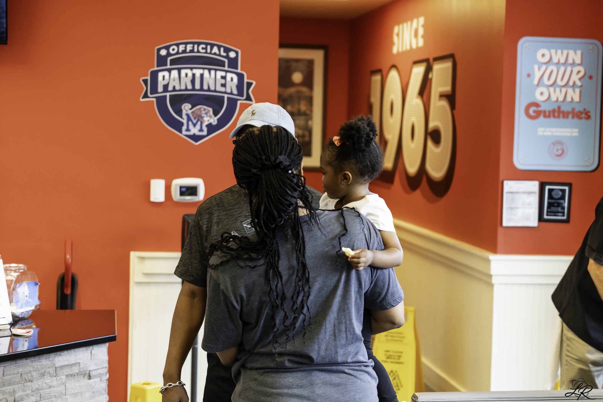 Two women and a young girl at the counter of a restaurant with red walls, a sign that says 'OFFICIAL PARTNER,' and promotional signage including 'Own Your Own Guthrie's.'