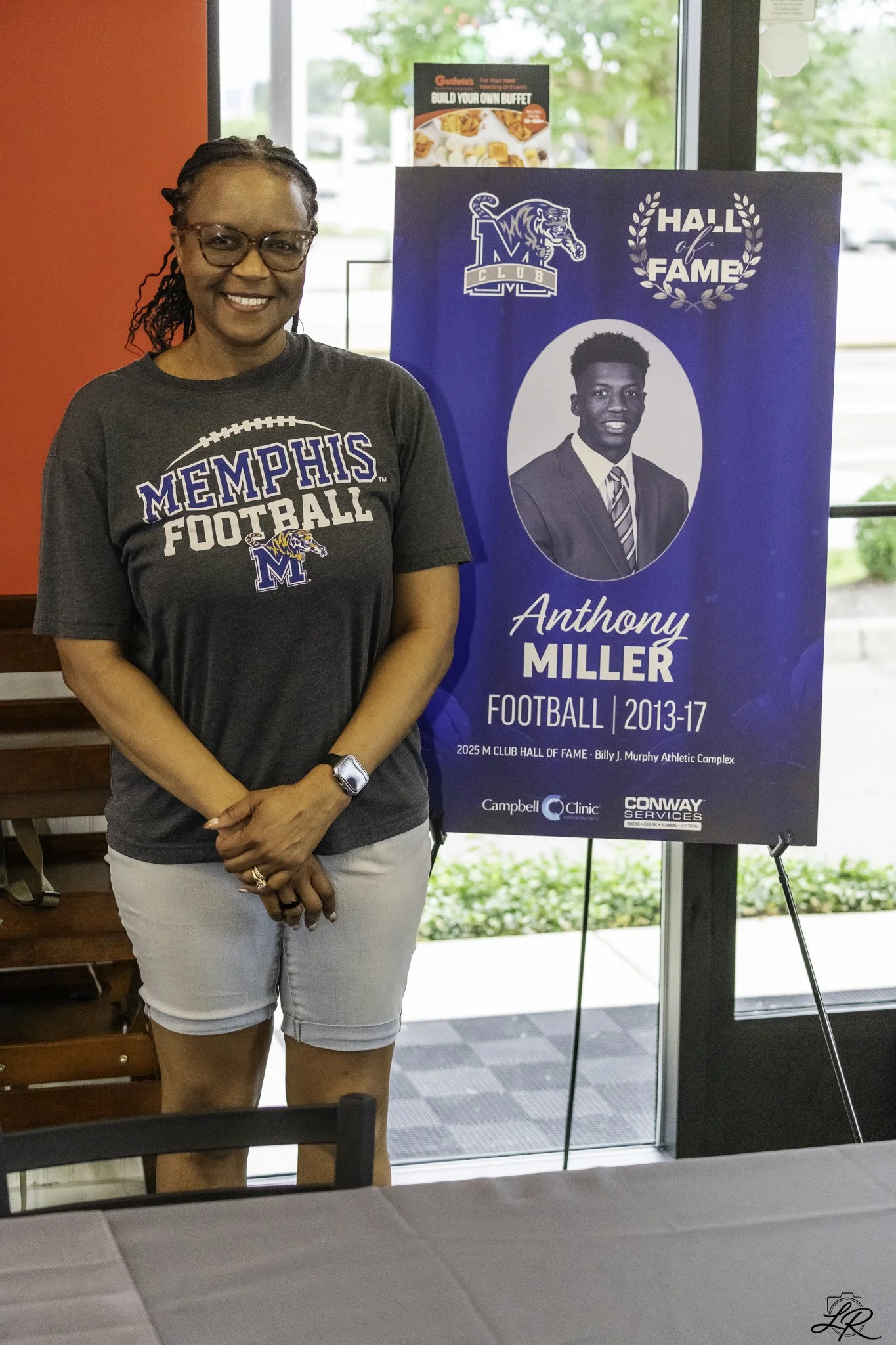 Woman standing next to a blue Hall of Fame sign, wearing a Memphis Football T-shirt, glasses, and a watch, smiling at the camera inside a building.