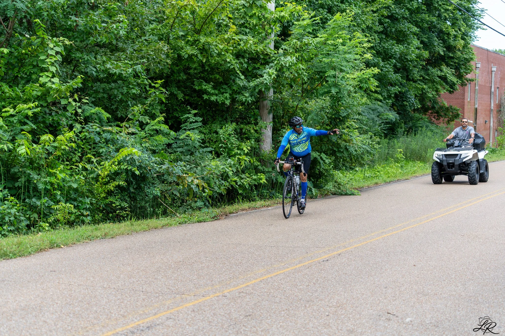 A man riding a bicycle on a road while another man on an ATV ride beside him, with lush green trees and a brick building in the background.