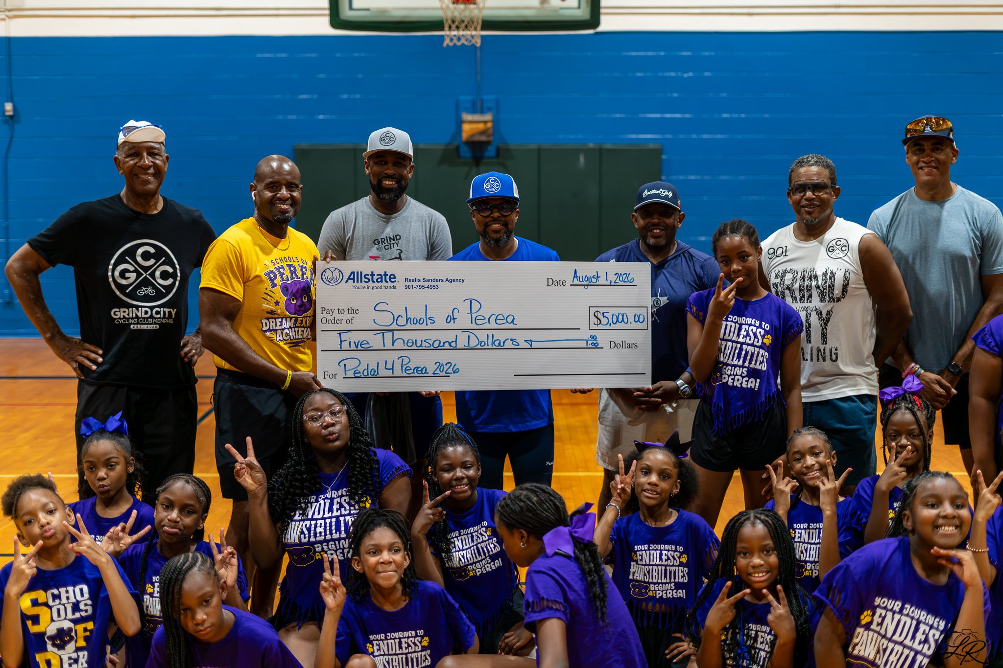 Group of children and adults posing for a photo in an indoor gymnasium. Adults hold a large check for $5,000 made out to Schools of Perea for Pedal 4 Perea 2026. The children wear matching blue T-shirts with yellow and purple text.