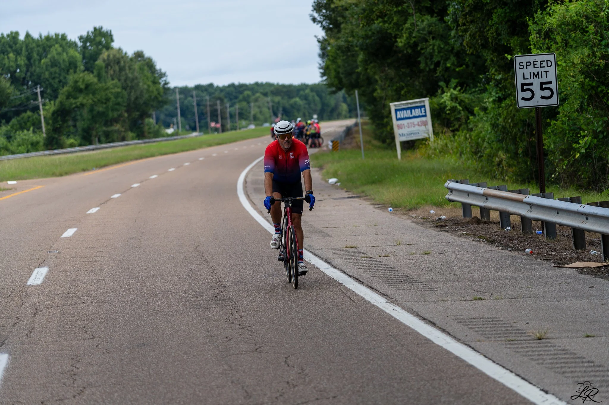 A cyclist wearing a helmet and sunglasses rides along the shoulder of a rural road with a speed limit sign of 55 mph, lush green trees, and a group of cyclists in the background.