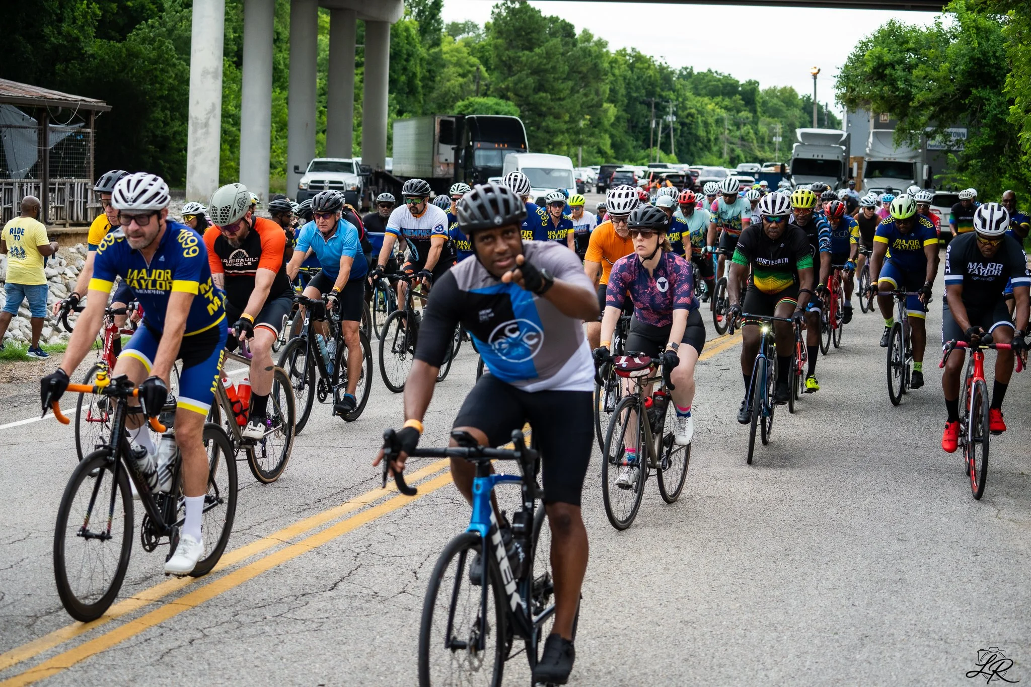 A large group of cyclists participating in a cycling event on a road with multiple cars and trucks parked or driving on the side. The cyclists are wearing helmets and cycling gear, with some people walking nearby.