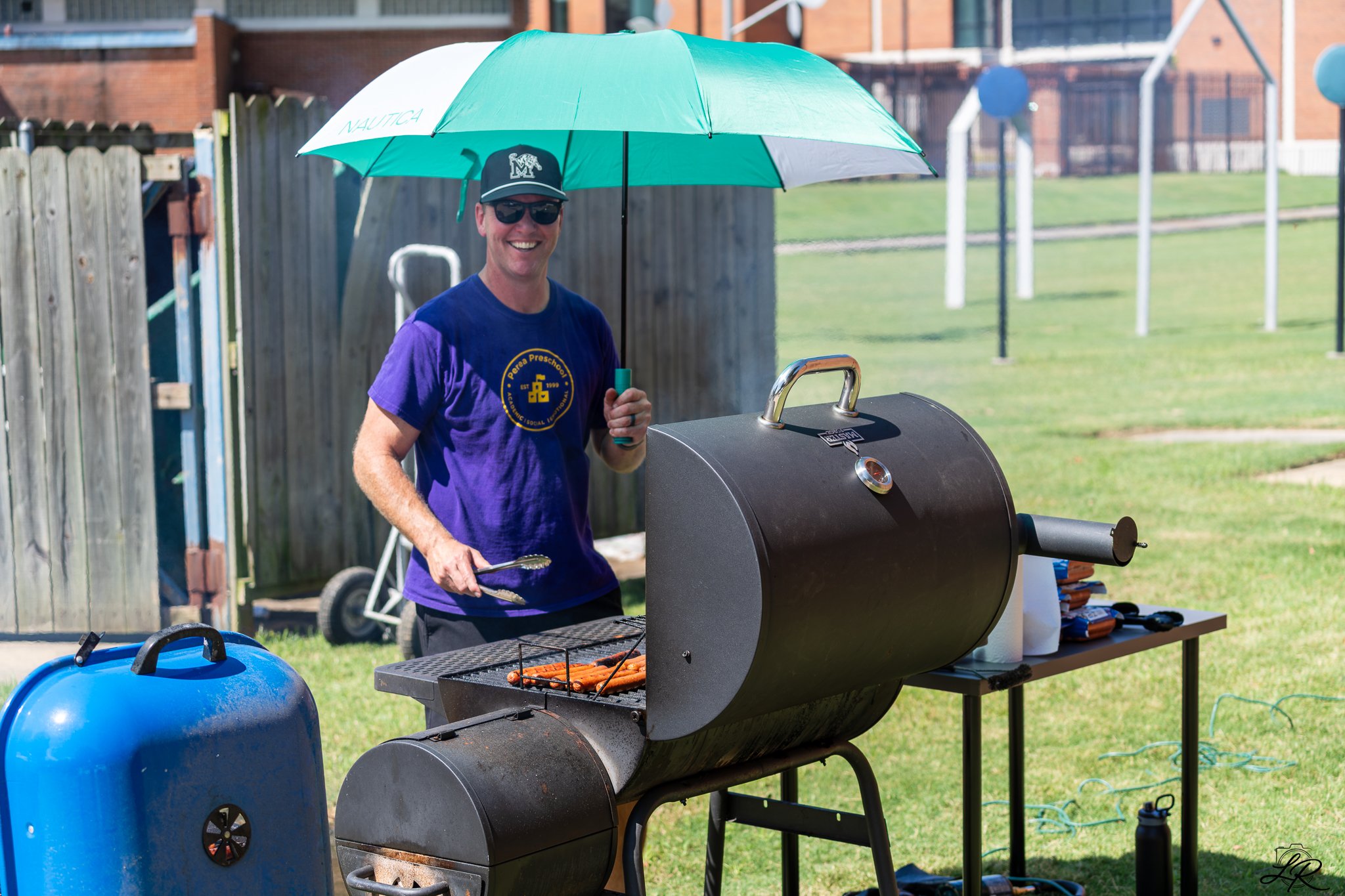 A smiling man wearing sunglasses, a baseball cap, and a blue T-shirt is standing next to a black charcoal grill outdoors. He is holding tongs and grilling hot dogs. The man is under a large green umbrella on a grassy area, with a blue propane tank ne