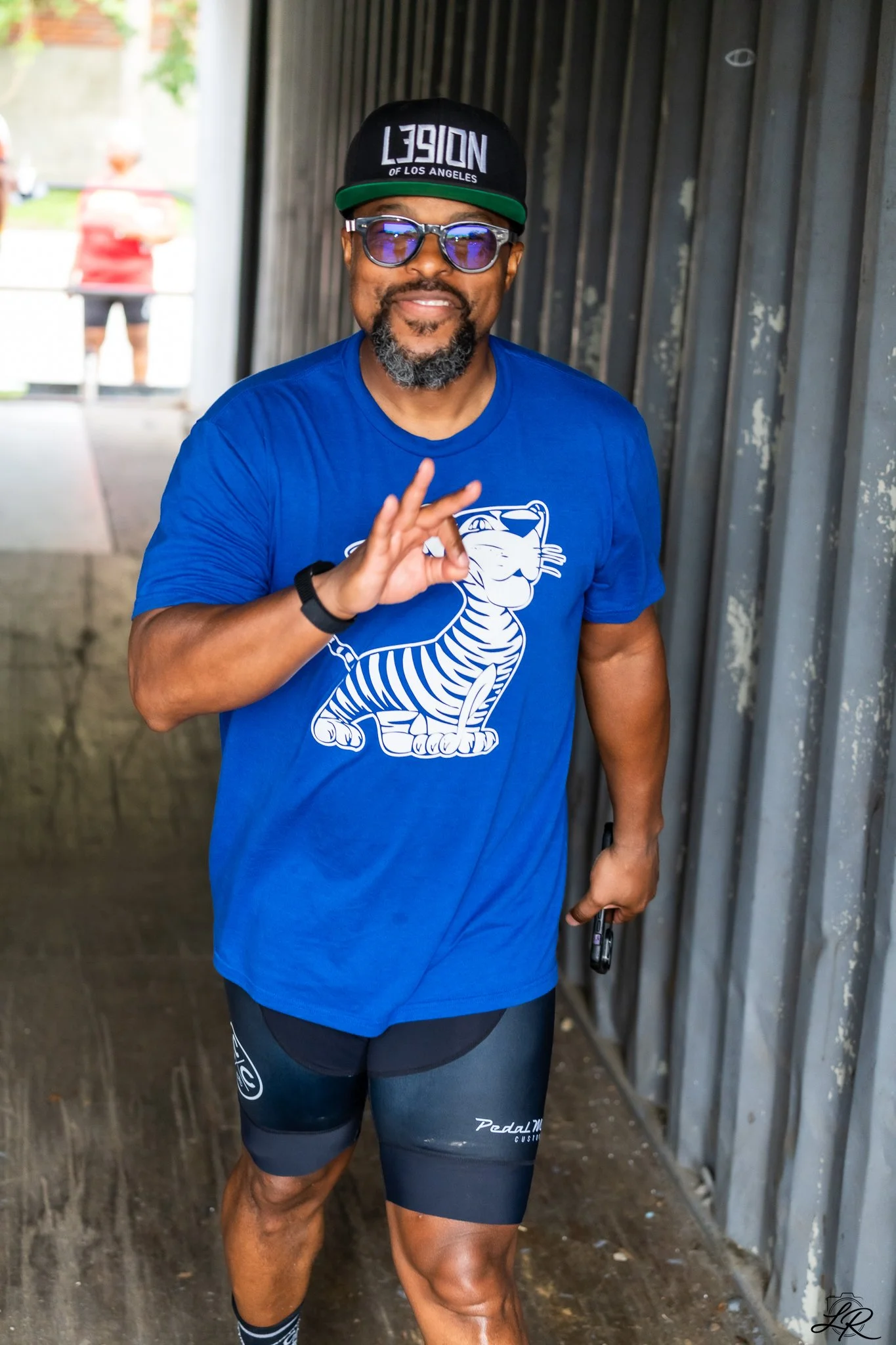 A man wearing sunglasses, a black cap with white text, a blue T-shirt with a tiger graphic, and black athletic shorts, smiling and making an ok hand gesture while standing at an outdoor location.