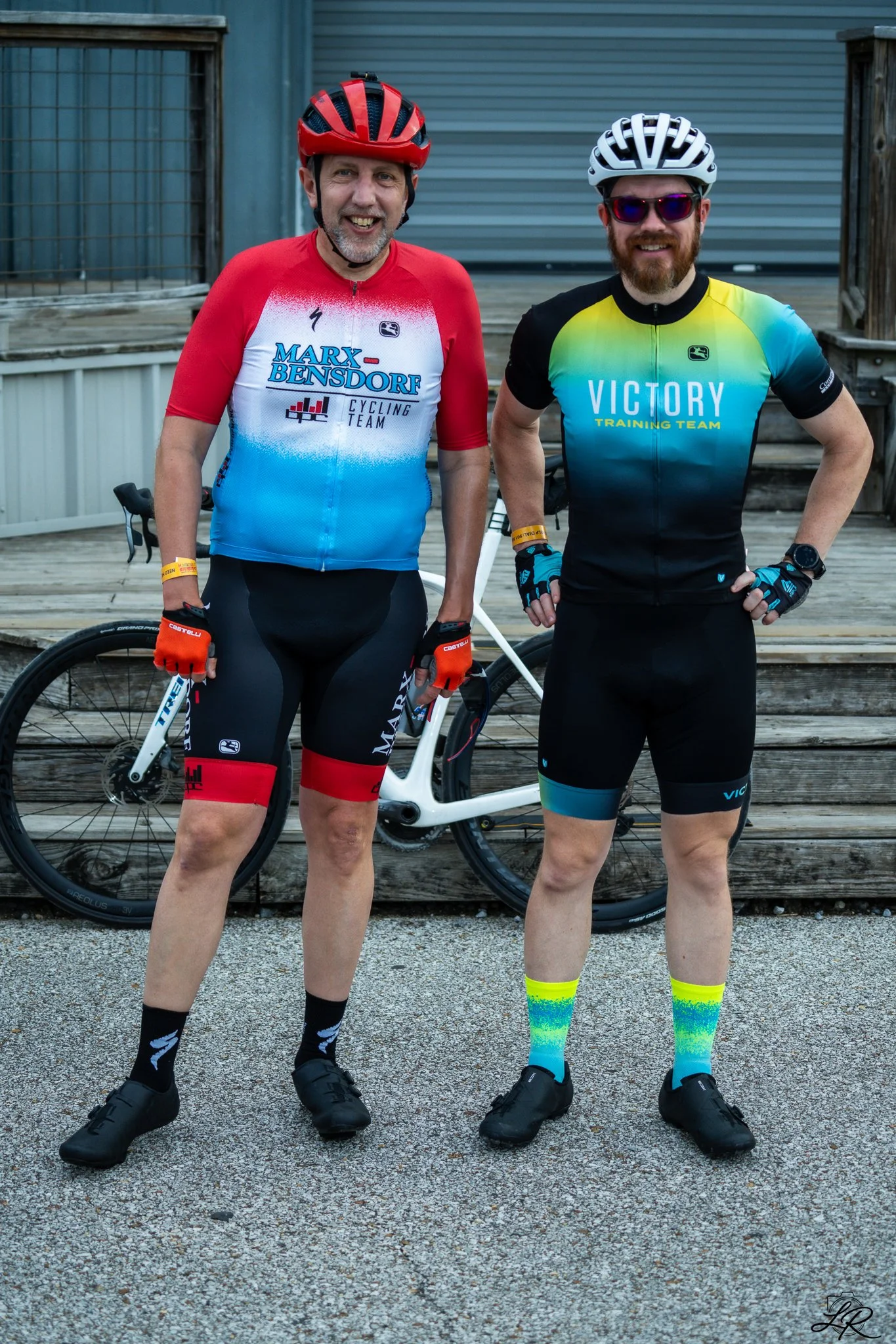 Two men in cycling gear standing outdoors with a bicycle, smiling, in front of a wooden deck and metal building. One wears a red, white, and blue jersey with 'Marx Bendorf Cycling Team' printed on it. The other wears a gradient yellow, green, and blu