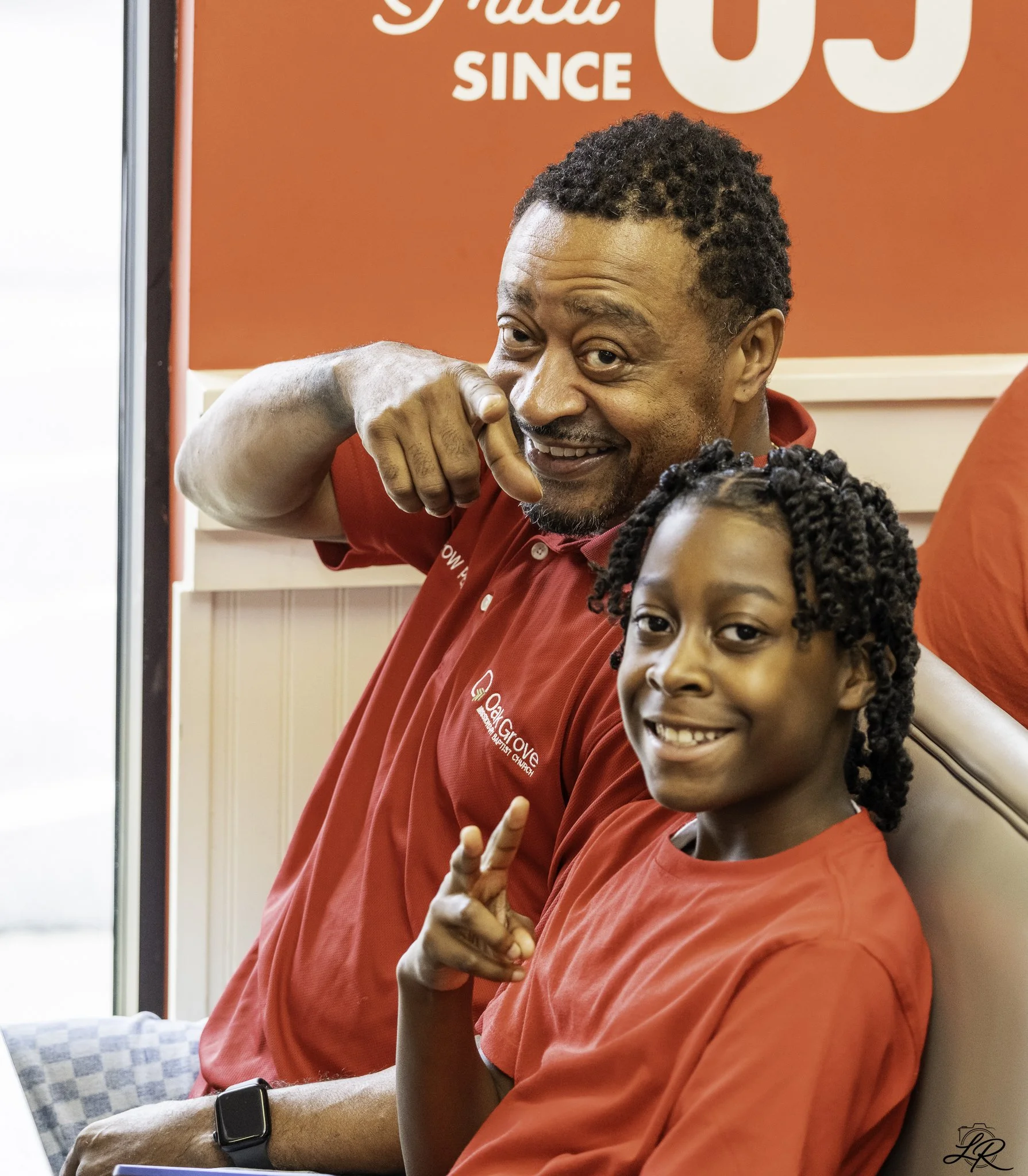 A man wearing a red shirt and a black wristwatch is smiling and making a peace sign with his right hand. There is a young girl in the foreground, also smiling, with braided hair. The background features a red wall with white text that reads 'Since' a