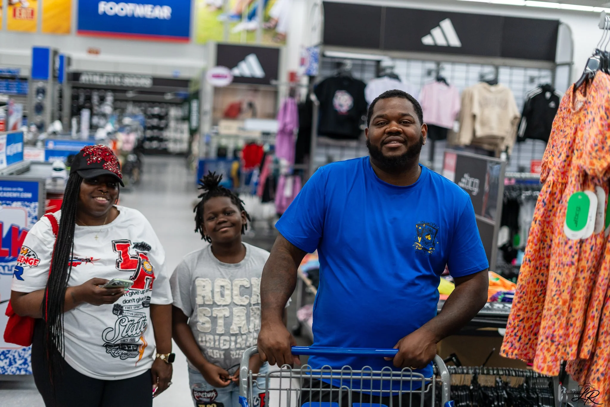 A group of diverse people at a retail store, with a man in a blue shirt at the center pushing a shopping cart, and a woman with braided hair wearing a white shirt with newsprint design, a young girl, and a woman with colorful dress nearby, surrounded