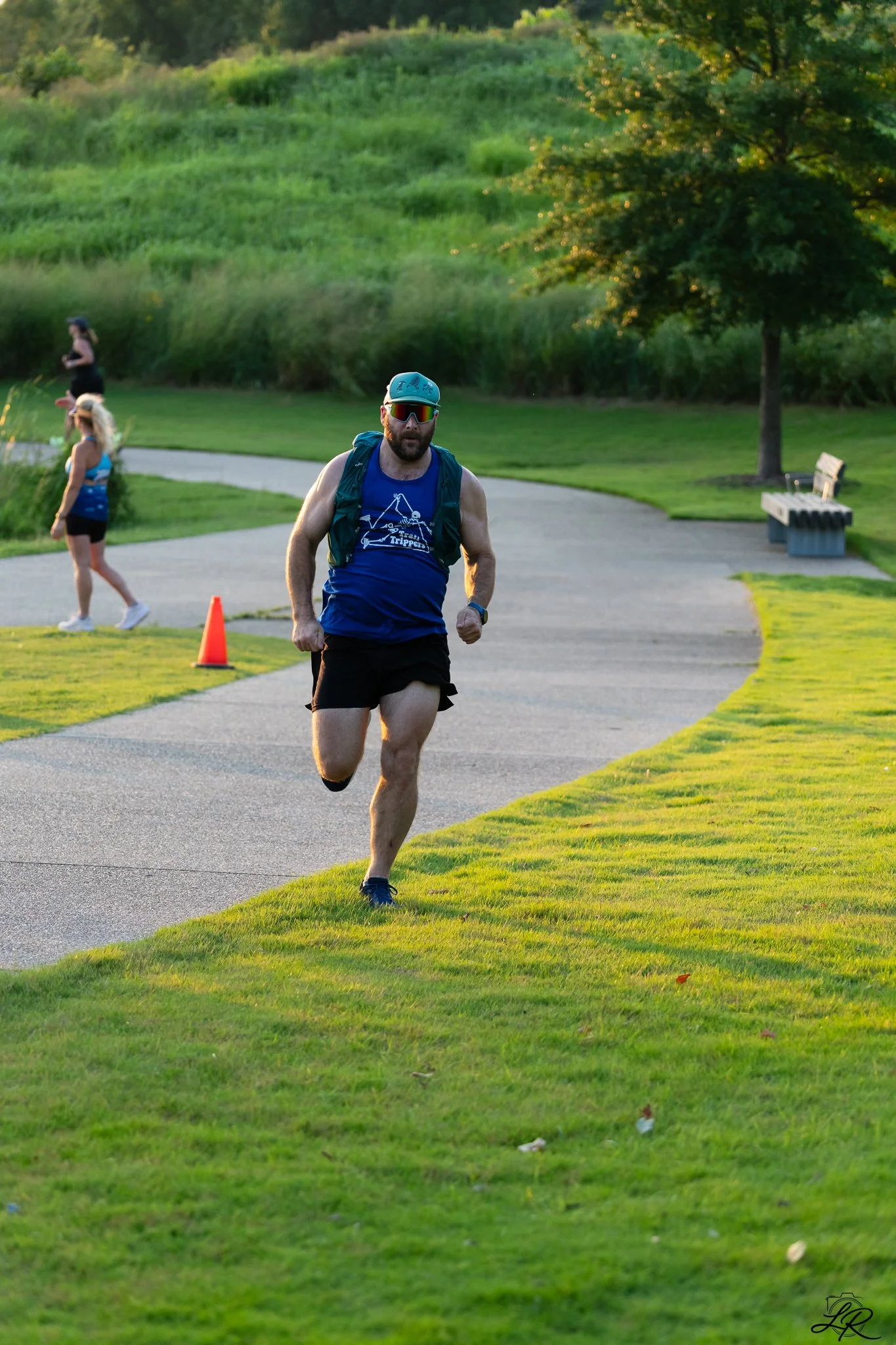 Man jogging on a park path with a grass field and hill in the background, accompanied by two women walking, one of whom is wearing a blue tank top and shorts while the other is running and the man is wearing sunglasses, a hat, and a sleeveless shirt.