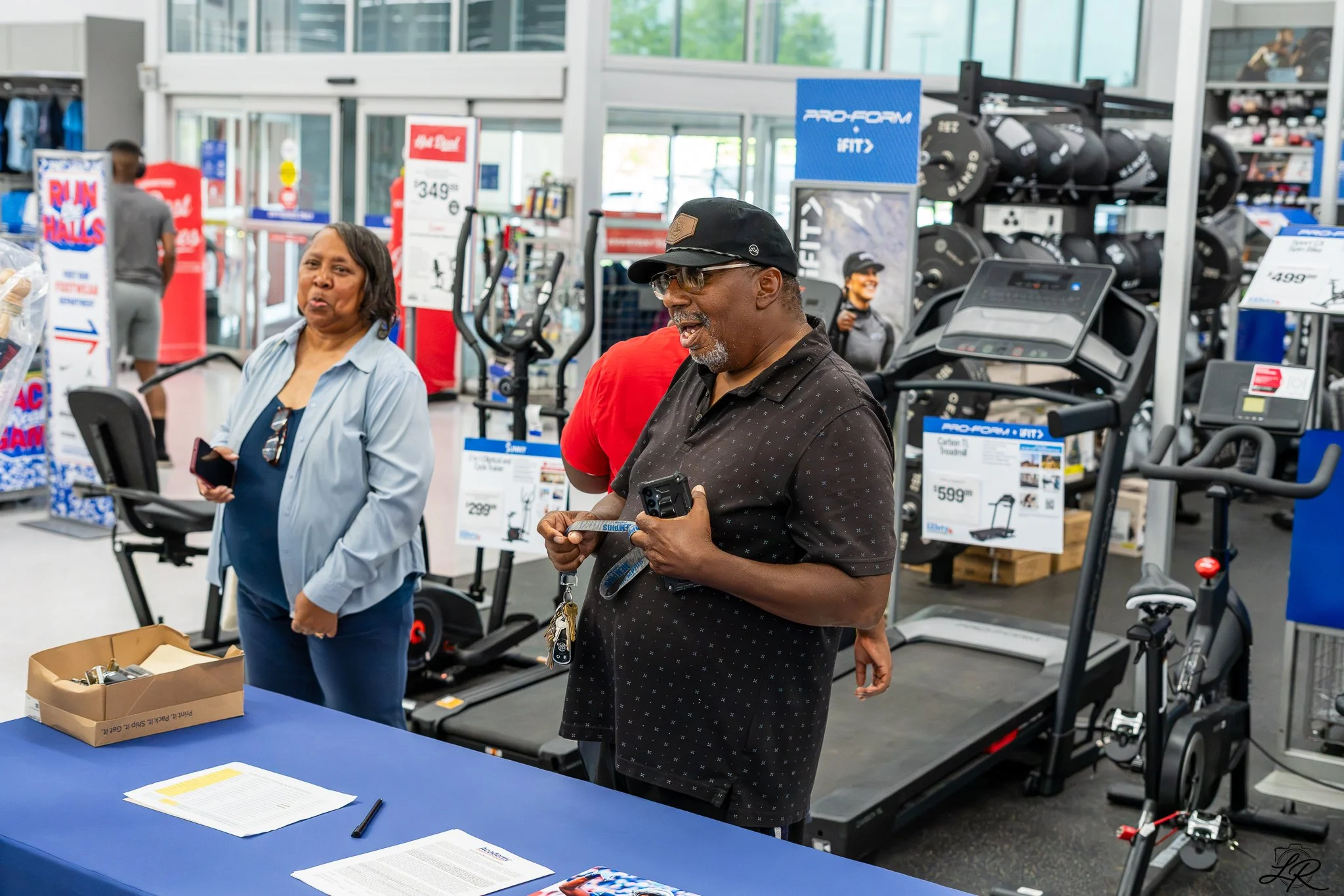 Two people are standing at a table inside a retail store that sells fitness equipment. The man on the right is wearing a black cap, glasses, and a black polo shirt, holding a pair of keys and smiling. The woman on the left, wearing a light blue shirt