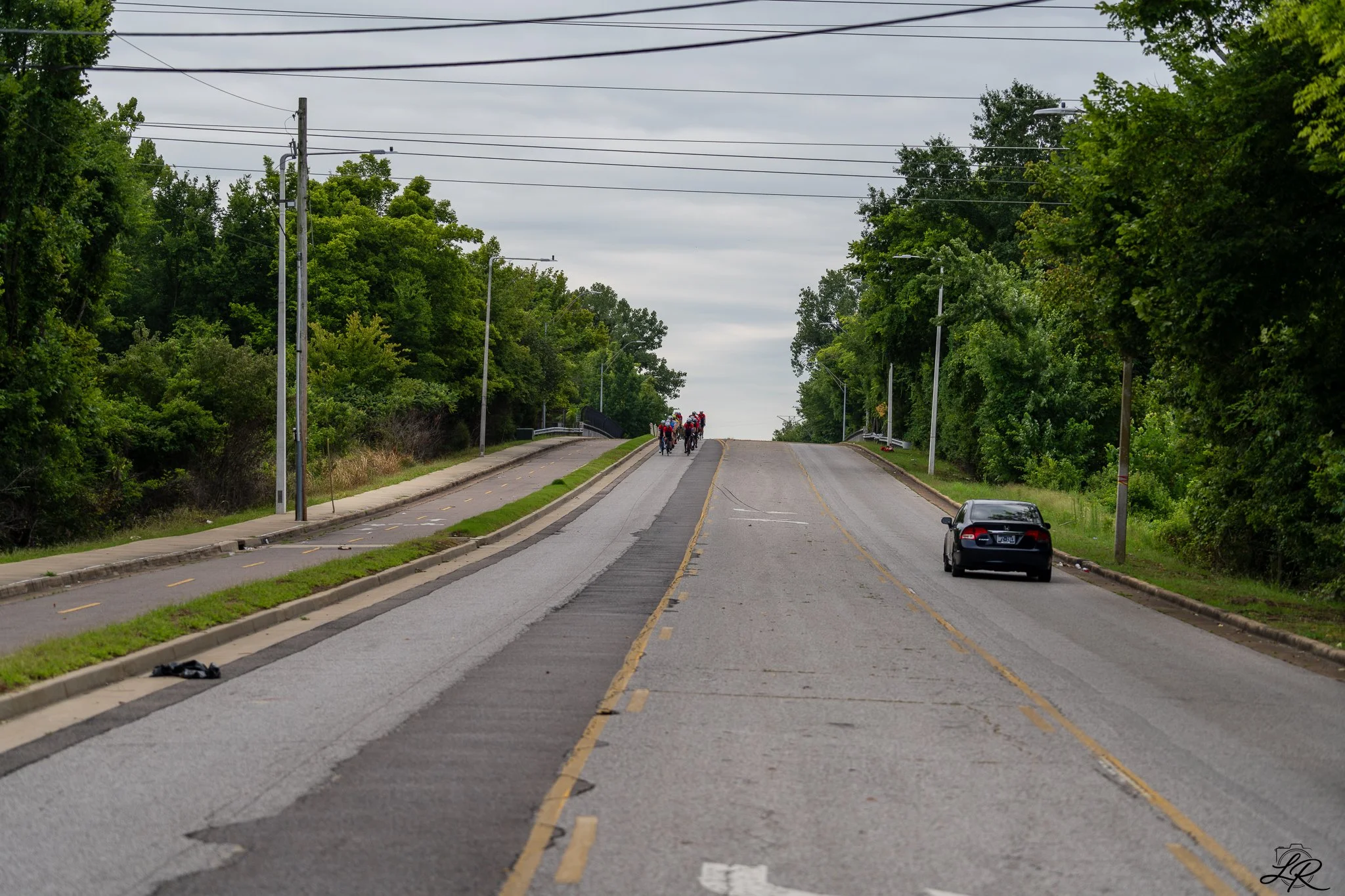 A street with a bike lane on the left, trees lining both sides, overcast sky, and a group of cyclists riding in the distance. A black car is parked on the right side.
