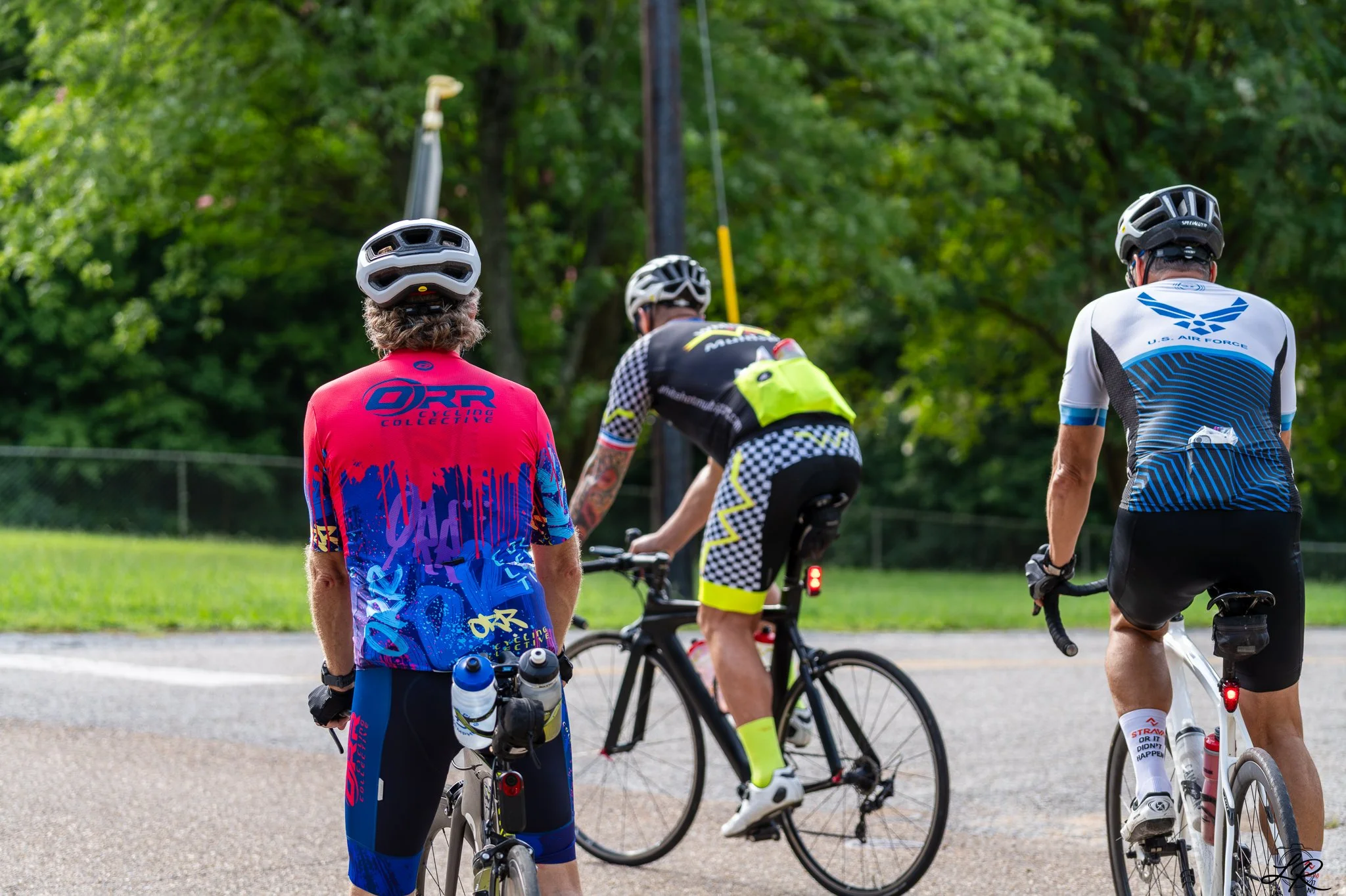 A group of cyclists, wearing helmets and colorful jerseys, gathered outdoors on a green area with trees in the background.