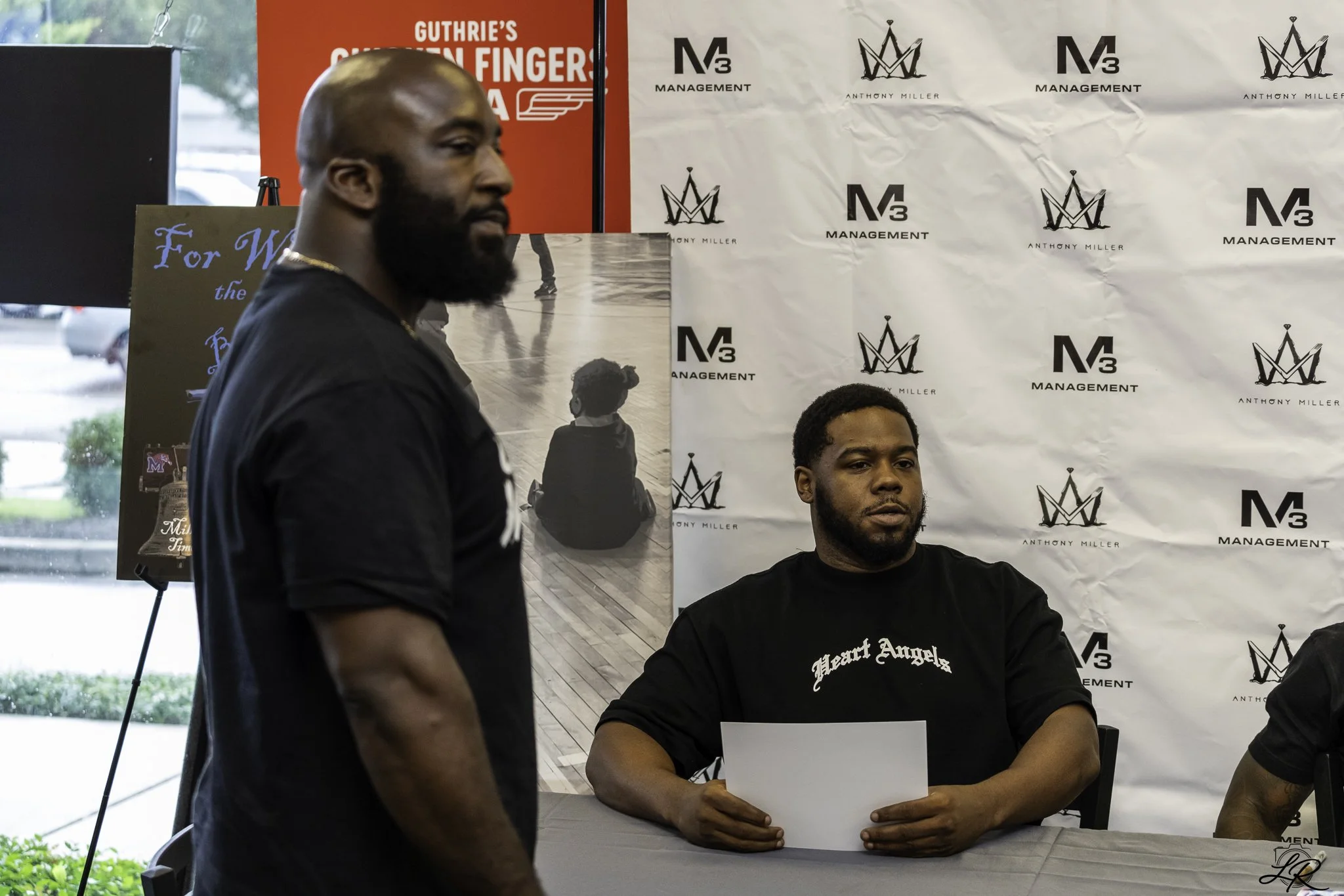 Two men are sitting at a table, one holding a paper, with a backdrop displaying logos and the words 'Anthony Miller' and 'M3 Management'. Another man stands nearby with a shaved head and beard, wearing a dark shirt and gold chain. In the background, 