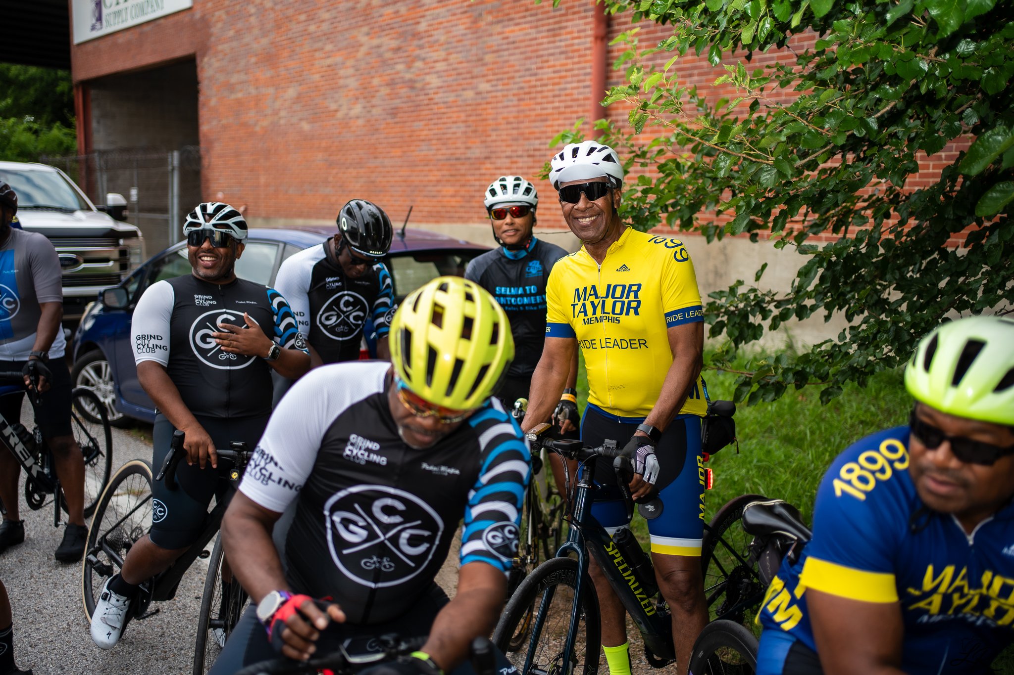 A group of cyclists gathered outdoors near a brick building, smiling and wearing cycling gear, helmets, and sunglasses.