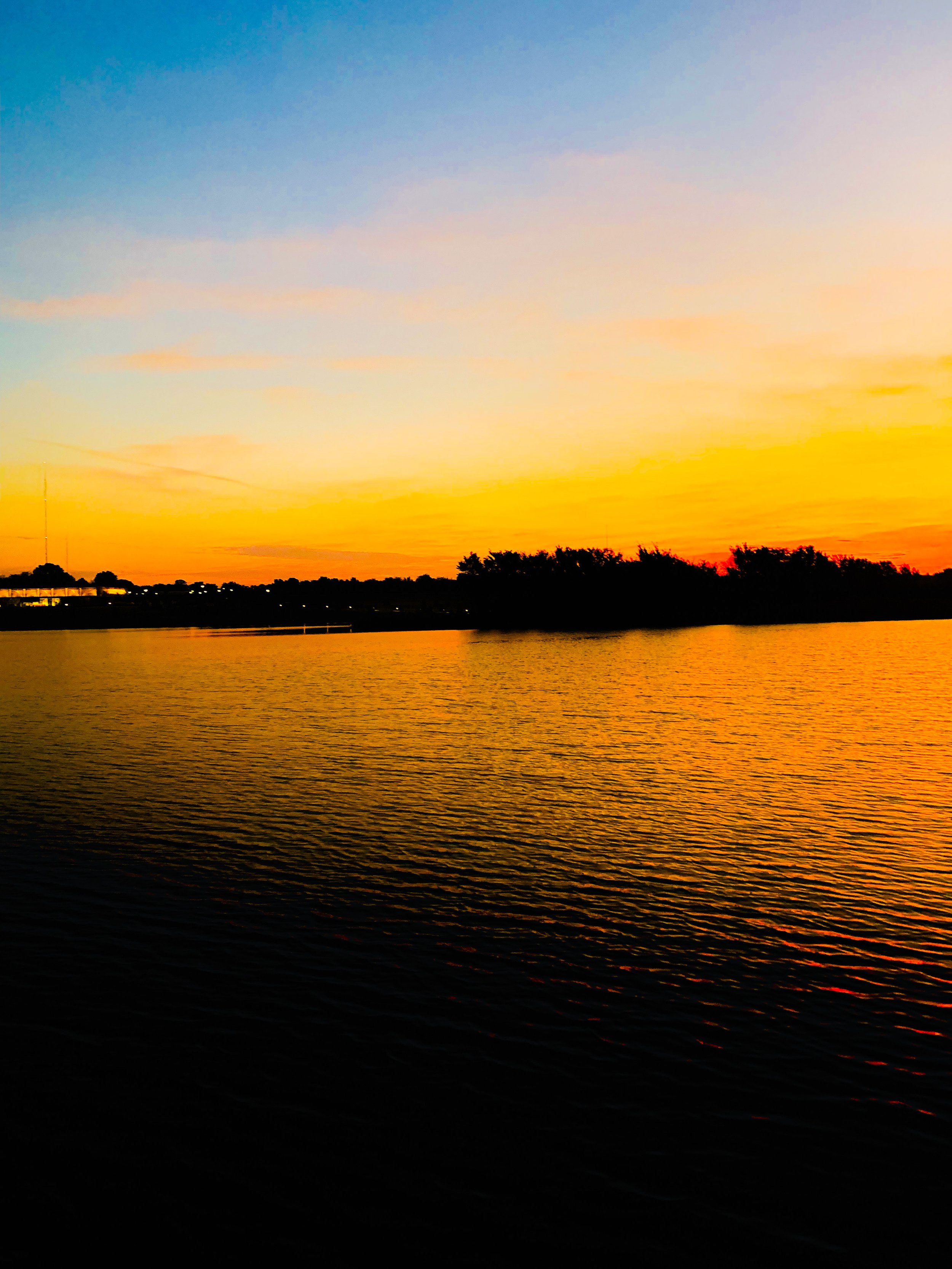 Sunset over a calm body of water with silhouettes of trees and a partly clear sky with shades of orange, yellow, and blue.