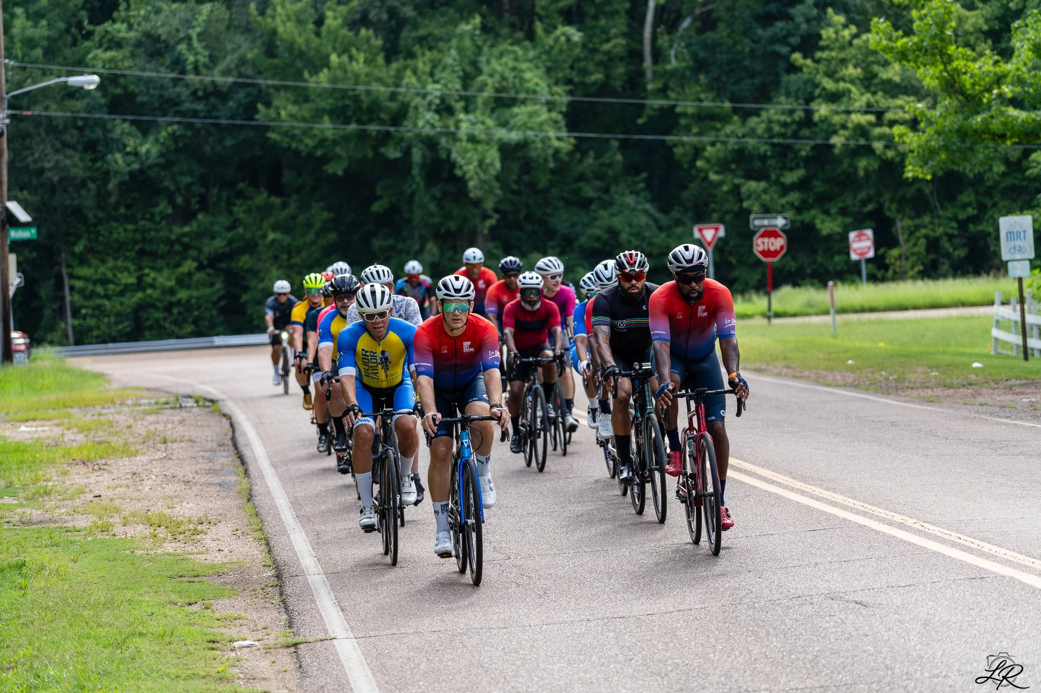 A group of cyclists riding on a two-lane road surrounded by greenery and trees, with multiple traffic signs including stop, yield, and bicycle signs visible in the background.