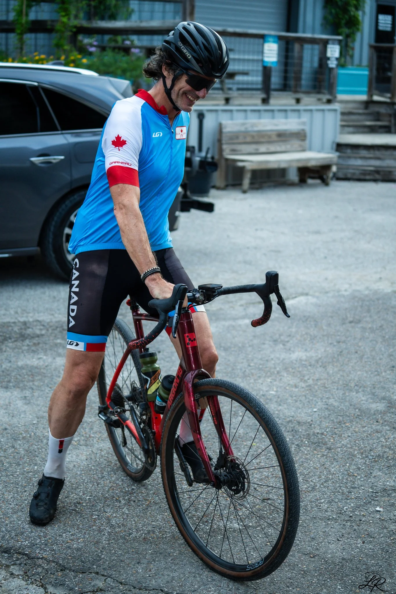 Cyclist wearing a Canadian cycling uniform, helmet, and sunglasses, standing with a red mountain bike on a paved outdoor area with benches and a building in the background.