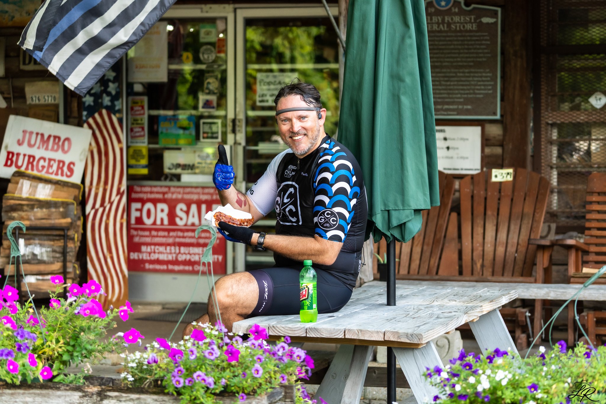 A man sitting on a picnic table in front of a store, holding a slice of pizza and smiling at the camera, with a green water bottle and a drink bottle on the table. He is wearing a cycling outfit and a headband, with a thumbs-up gesture. There are pur