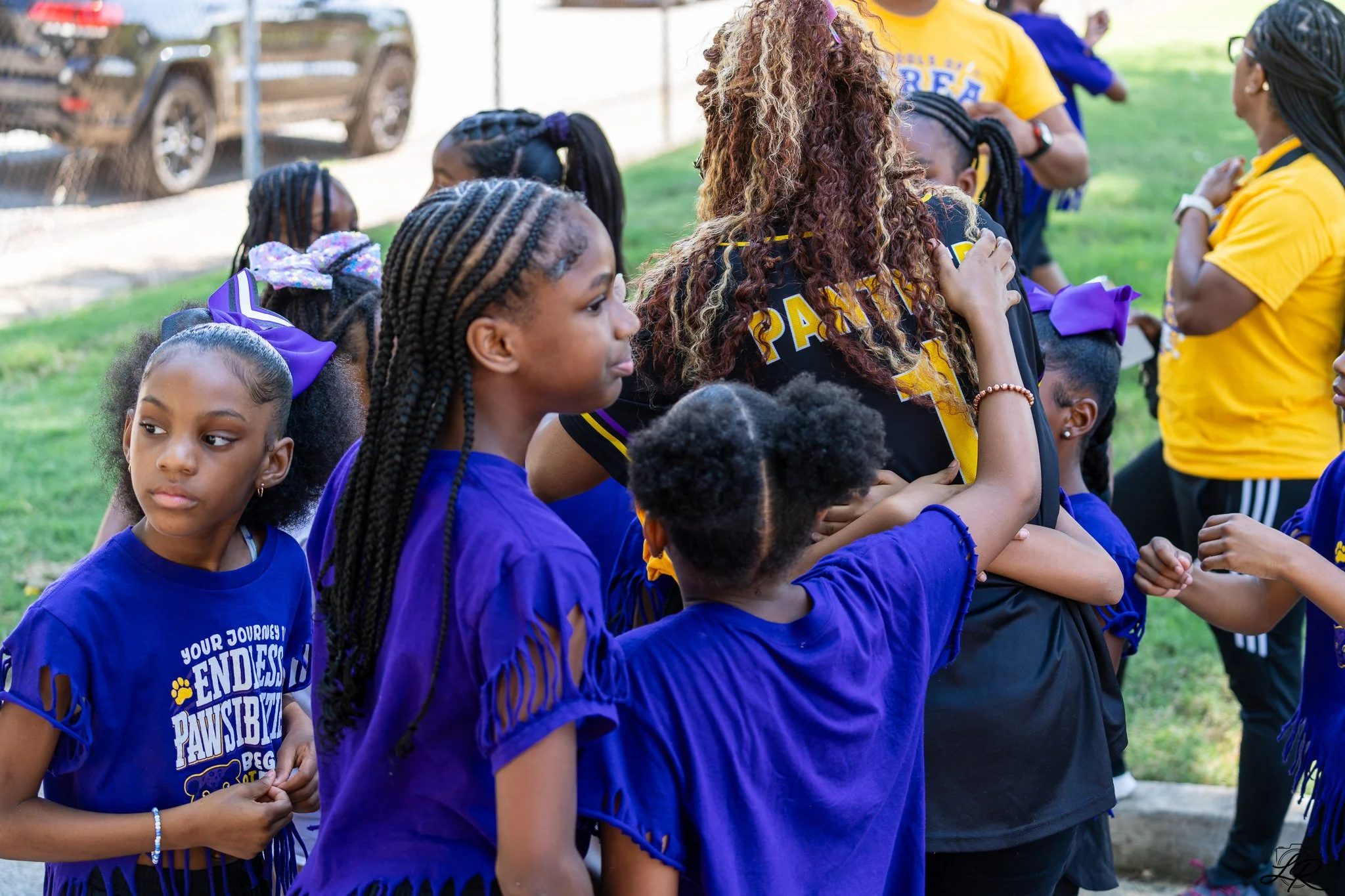 A group of young girls wearing blue shirts and purple bows, gathered outdoors. They are standing in a circle, participating in a group activity, with some adults in yellow shirts talking nearby.