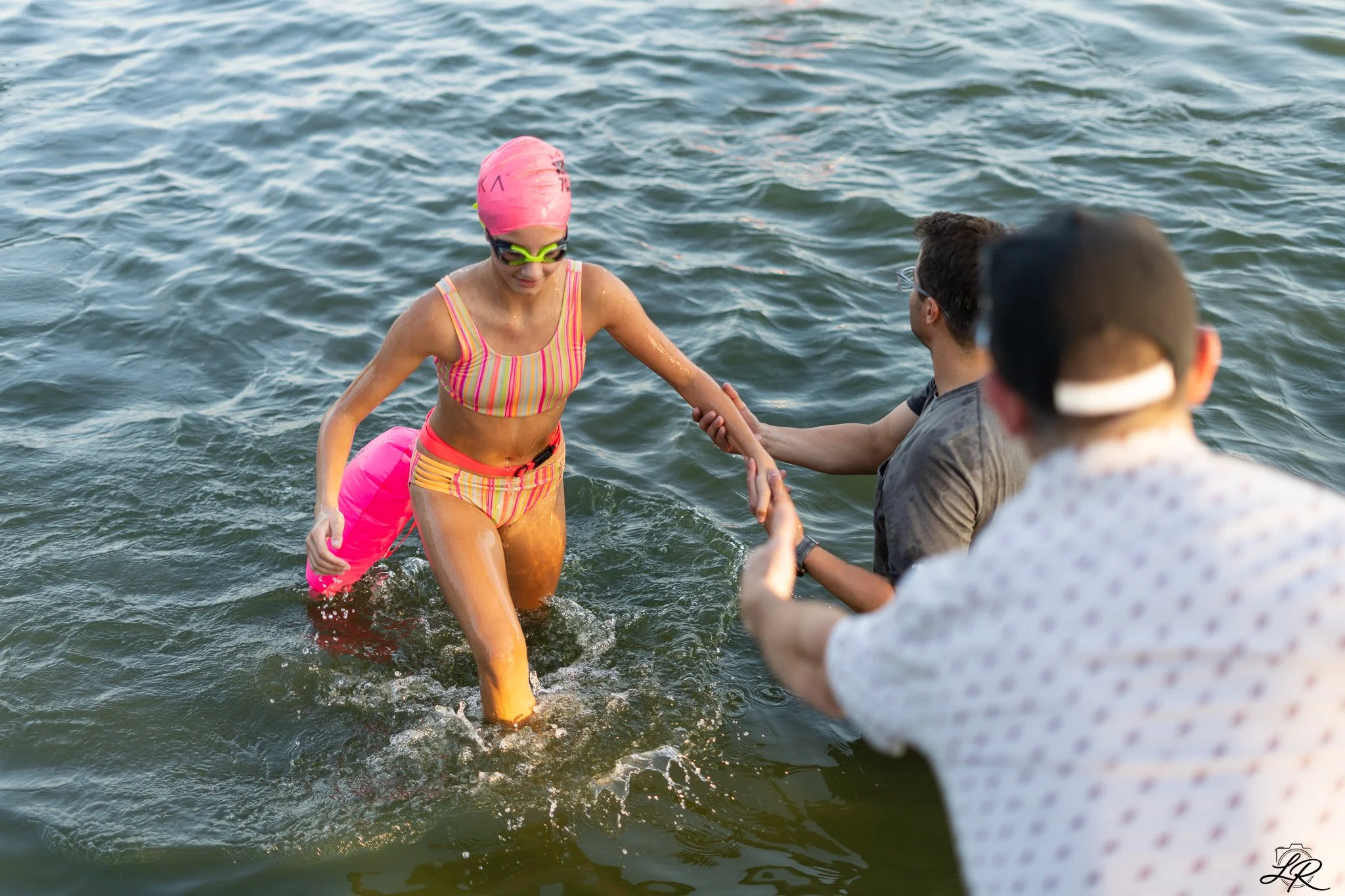 A woman in a colorful swimsuit, pink swim cap, and goggles emerging from a lake while holding hands with people on a boat or dock, with rippling water in the background.