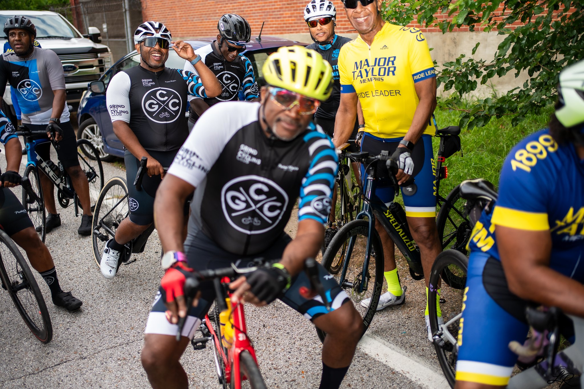 A group of cyclists dressed in cycling gear, some wearing helmets and sunglasses, standing together and smiling during a ride, with cars and a brick wall in the background.