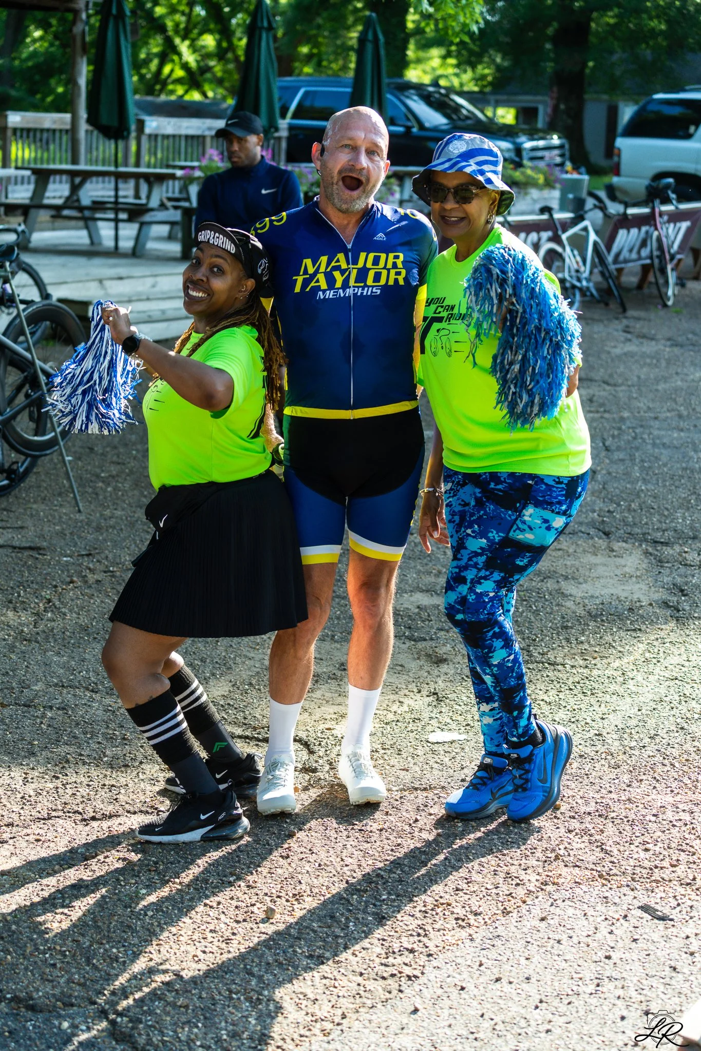 Three people standing outdoors, smiling and posing for a photo. The person in the middle is a man wearing a blue cycling jersey with yellow lettering that says 'Major Taylor Memphis', white cycling shoes, and white socks. The person on the left is a 