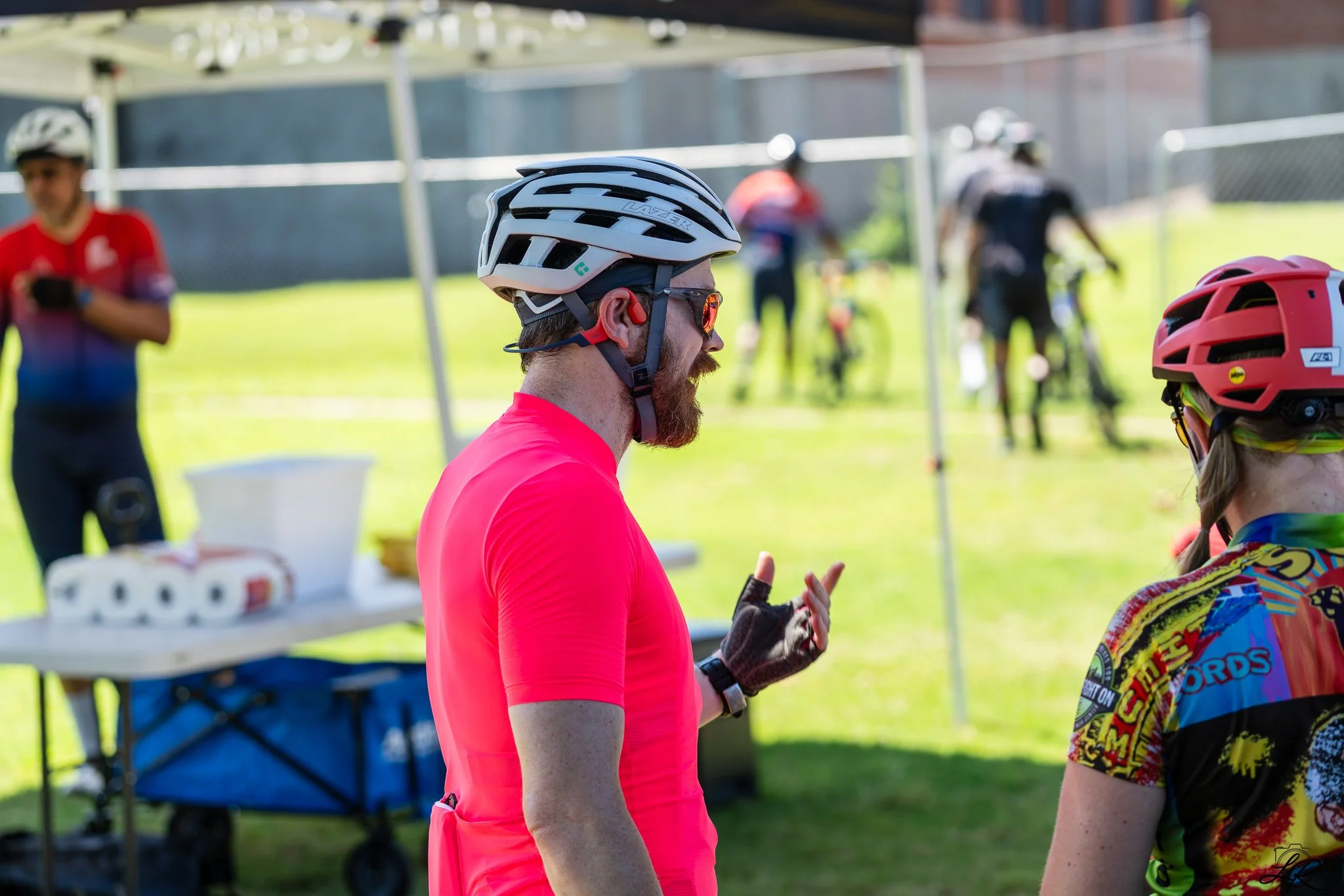 A group of people outdoors, some wearing helmets and cycling gear, engaging in conversation near a table with supplies.
