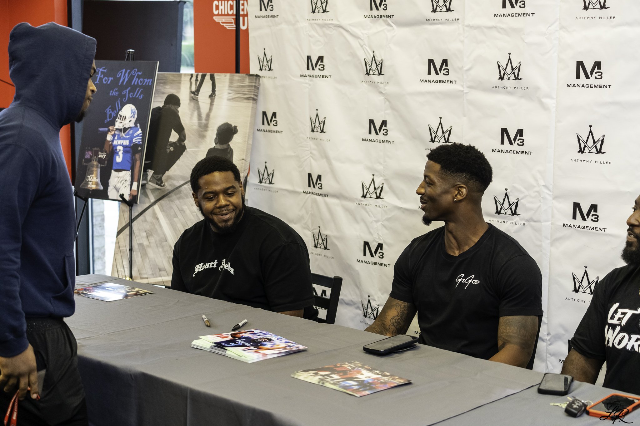 Three men sitting at a table during a signing event, with a man in a hoodie standing in front of them. The backdrop features logos for Anthony Miller and M3 Management. The table has promotional magazines and pens on it.