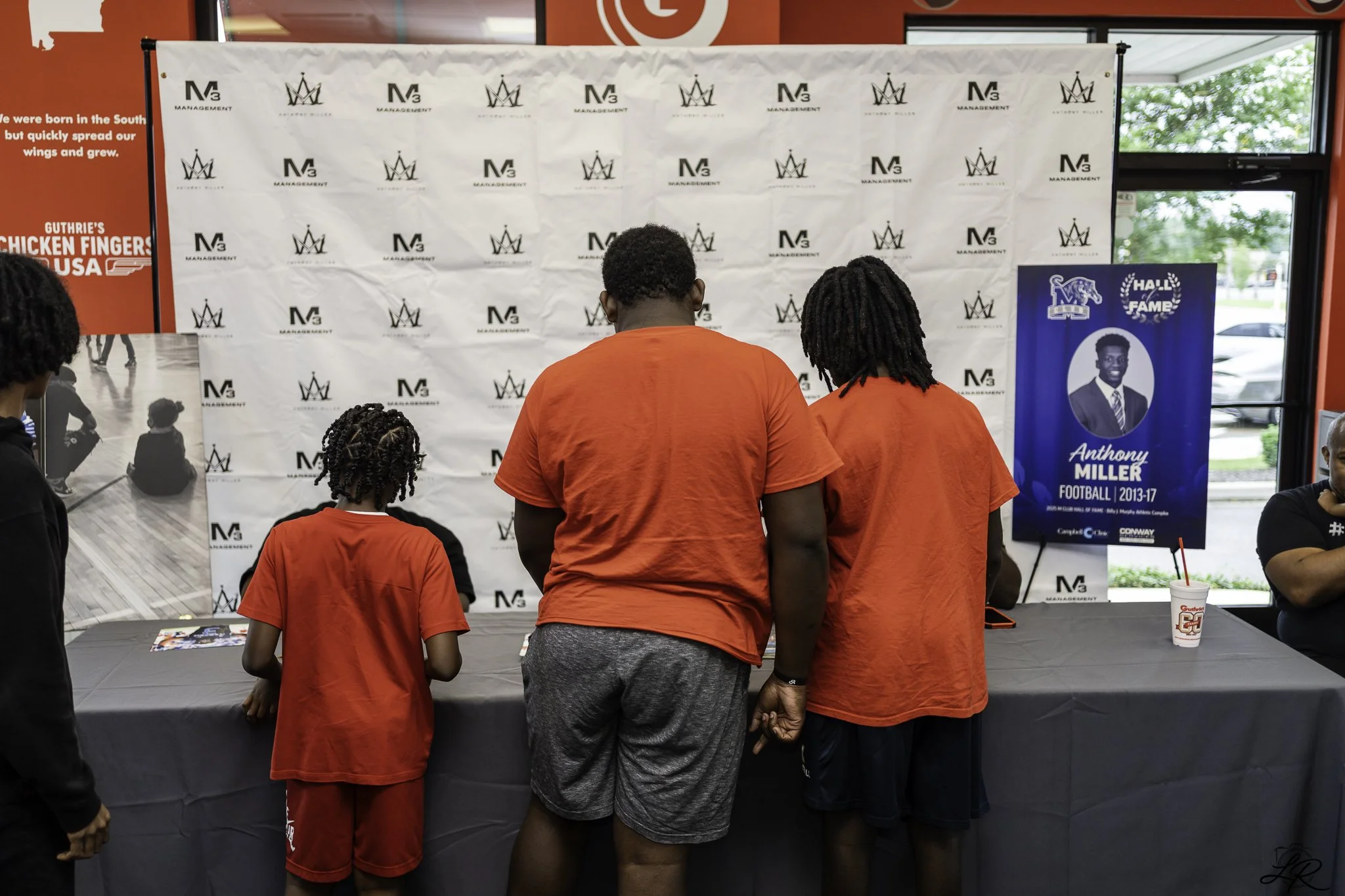 Four people, wearing red shirts, standing at a table with a black tablecloth, facing a white backdrop with logos. One person is a young girl and the other three are adult men. Behind them is a blue Hall of Fame sign with a picture of Anthony Miller, 