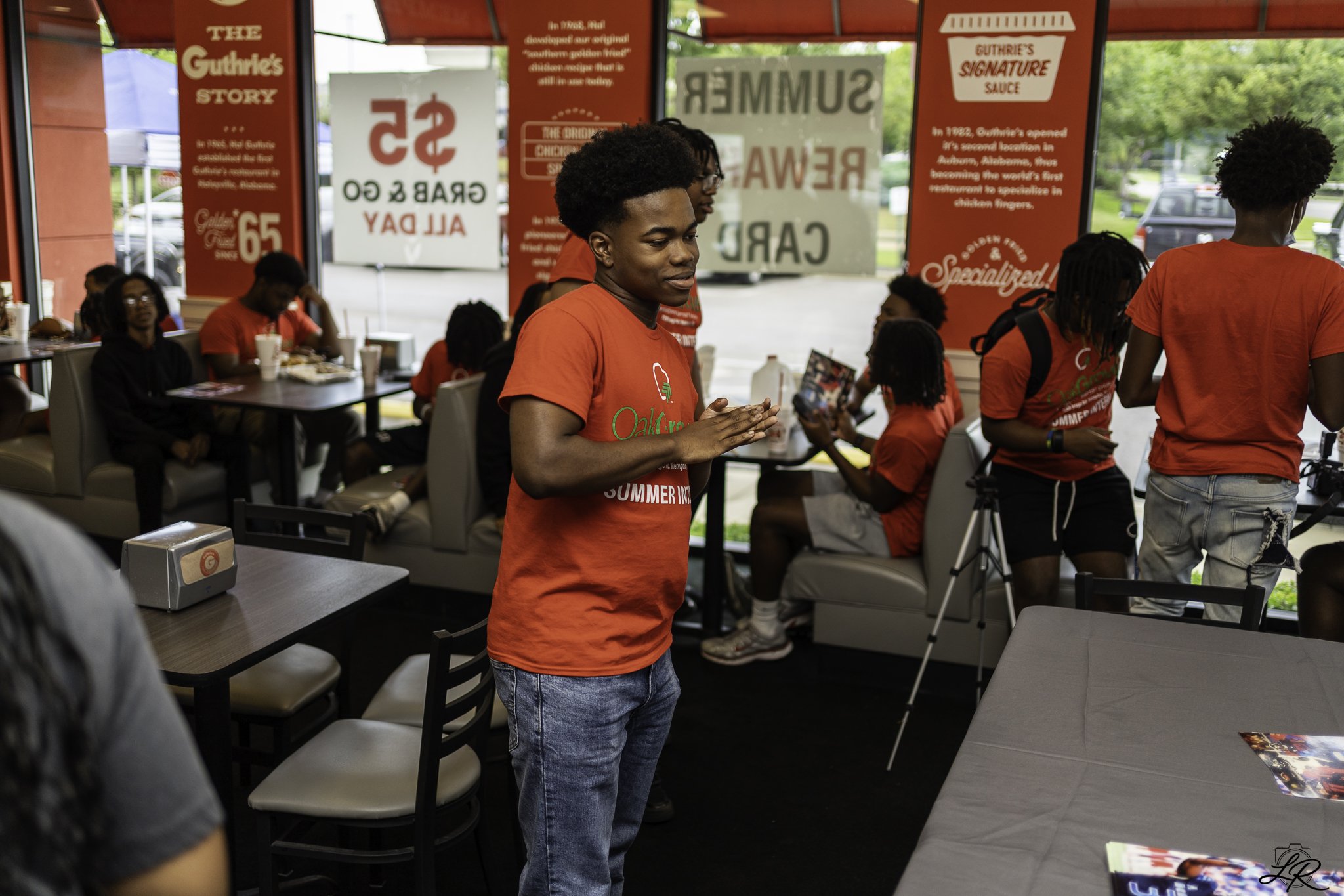 A group of people inside a fast-food restaurant, some sitting and some standing, with red-themed decor and signs on the walls, including a young man in a red t-shirt in the foreground