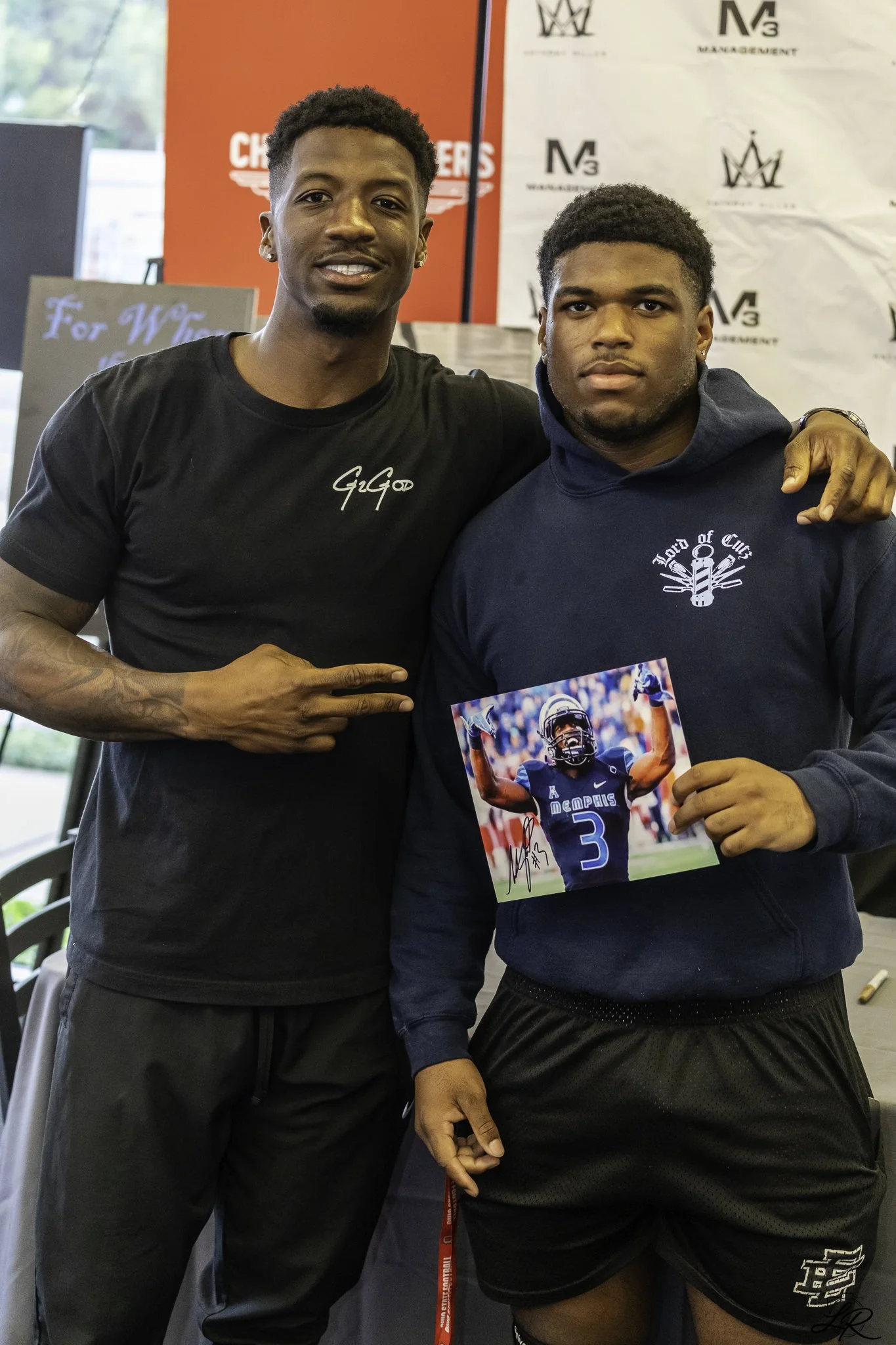 Two young men standing together indoors, one wearing a black t-shirt with the name 'Gigo' and the other in a navy hoodie holding a signed photo of a football player, with banners in the background.