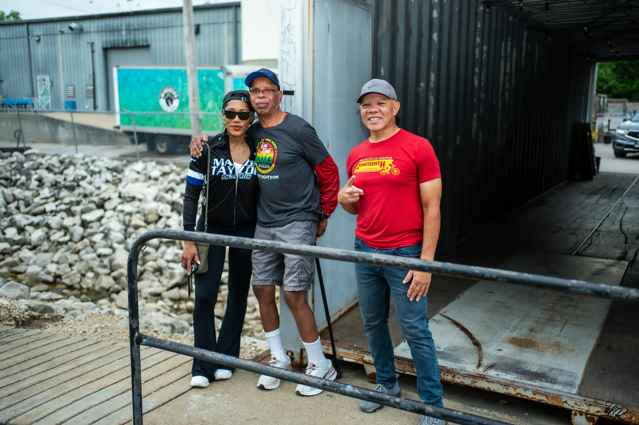 Three people standing together outdoors in front of a small train and rocks, posing for a photo. The woman on the left is wearing sunglasses, a black jacket, and dark pants; the man in the middle has glasses, a cap, and a dark T-shirt with a colorful