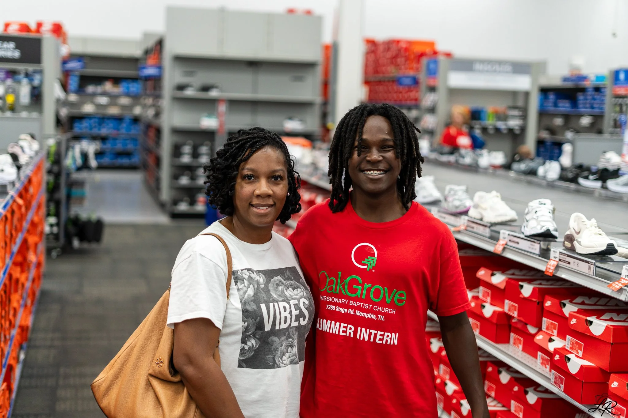 Two women standing inside a retail store in front of shoe racks, smiling at the camera. The woman on the left wears a white T-shirt with floral print and has a tan purse, while the woman on the right wears a red T-shirt with green text and a logo.