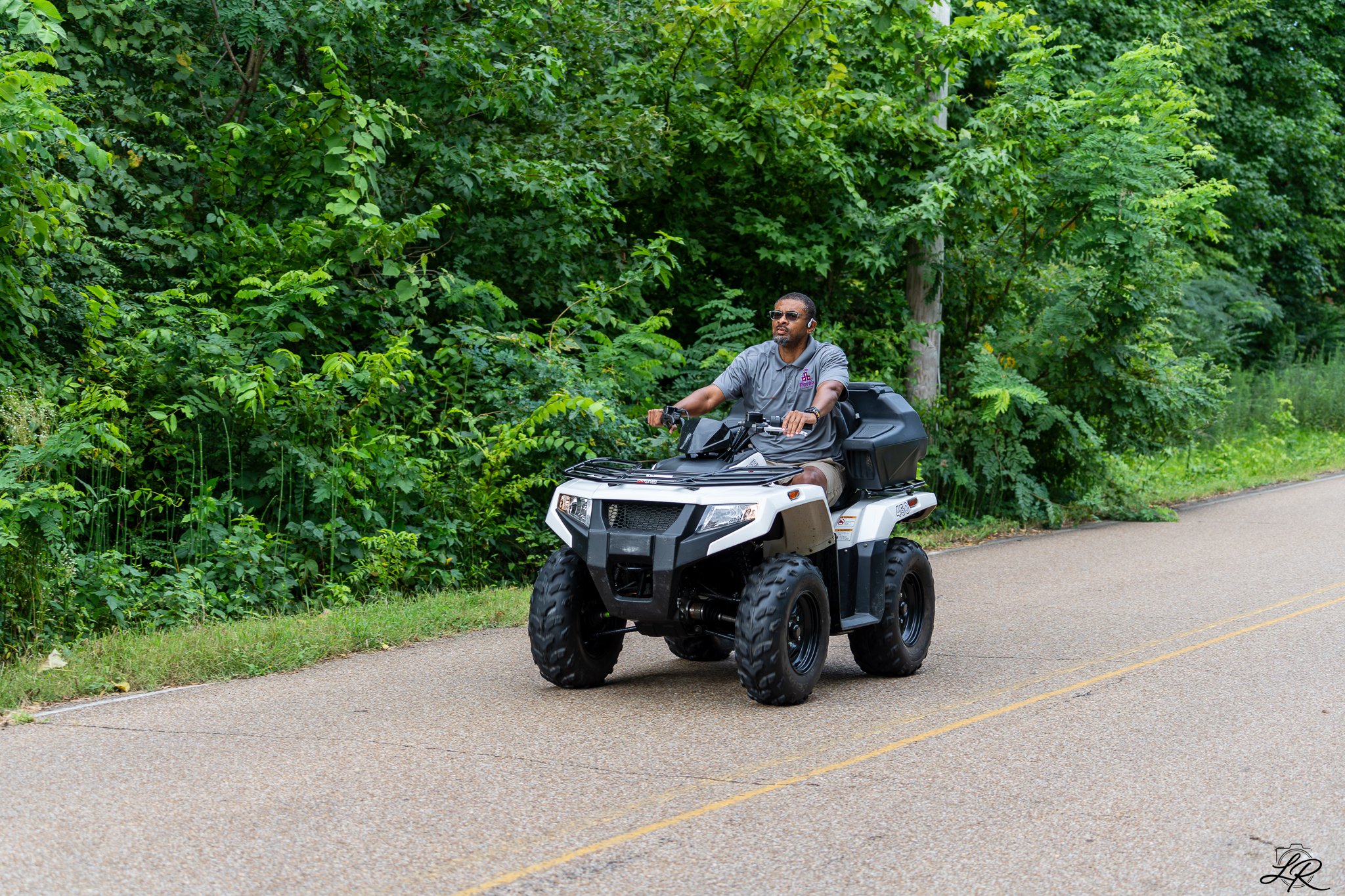 A man in sunglasses riding a black and white all-terrain vehicle (ATV) on a paved road next to dense green foliage.