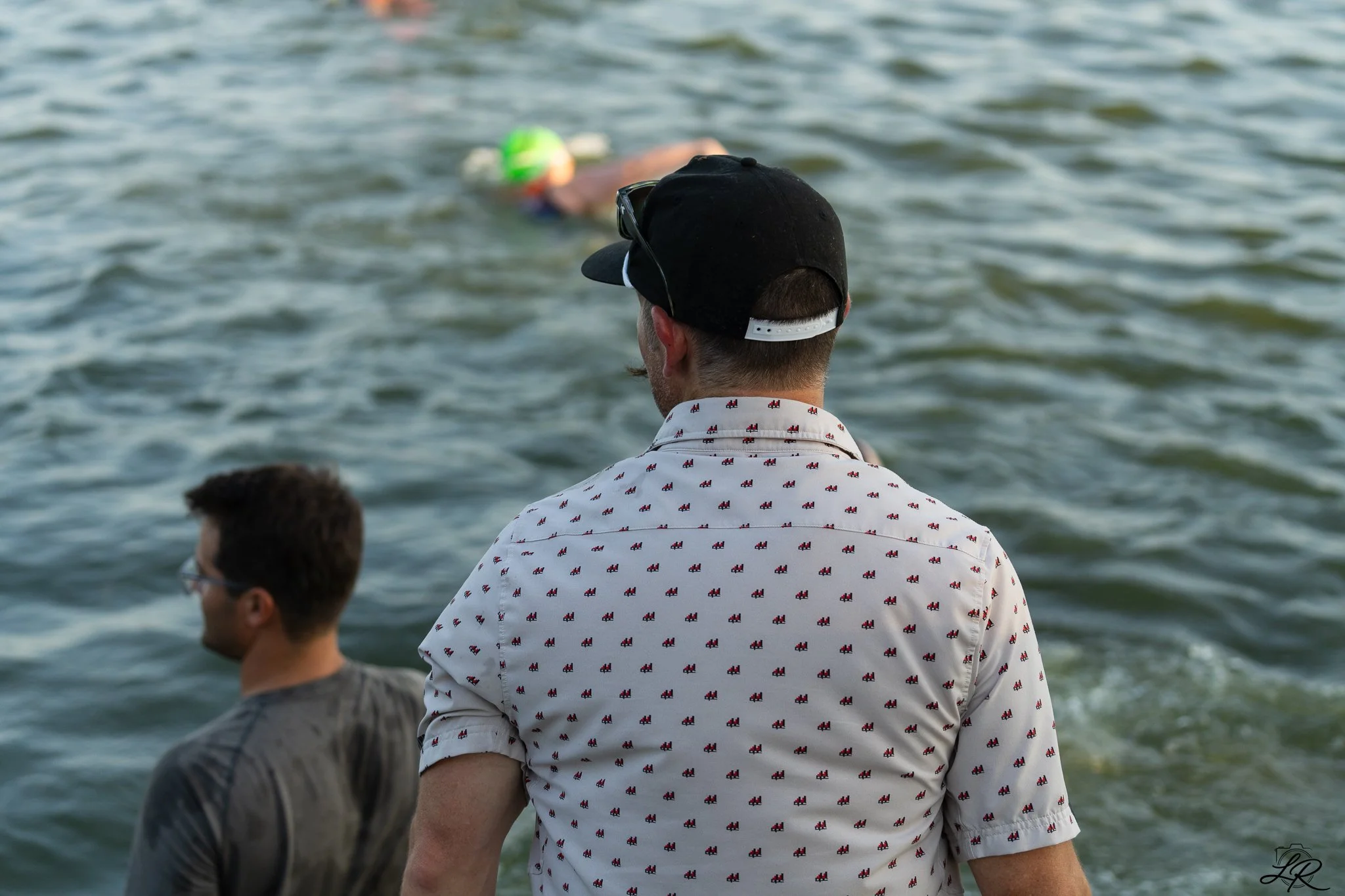 People standing near water, looking towards the water, with one person wearing a white shirt with red and blue patterns and a black cap, in an outdoor setting.