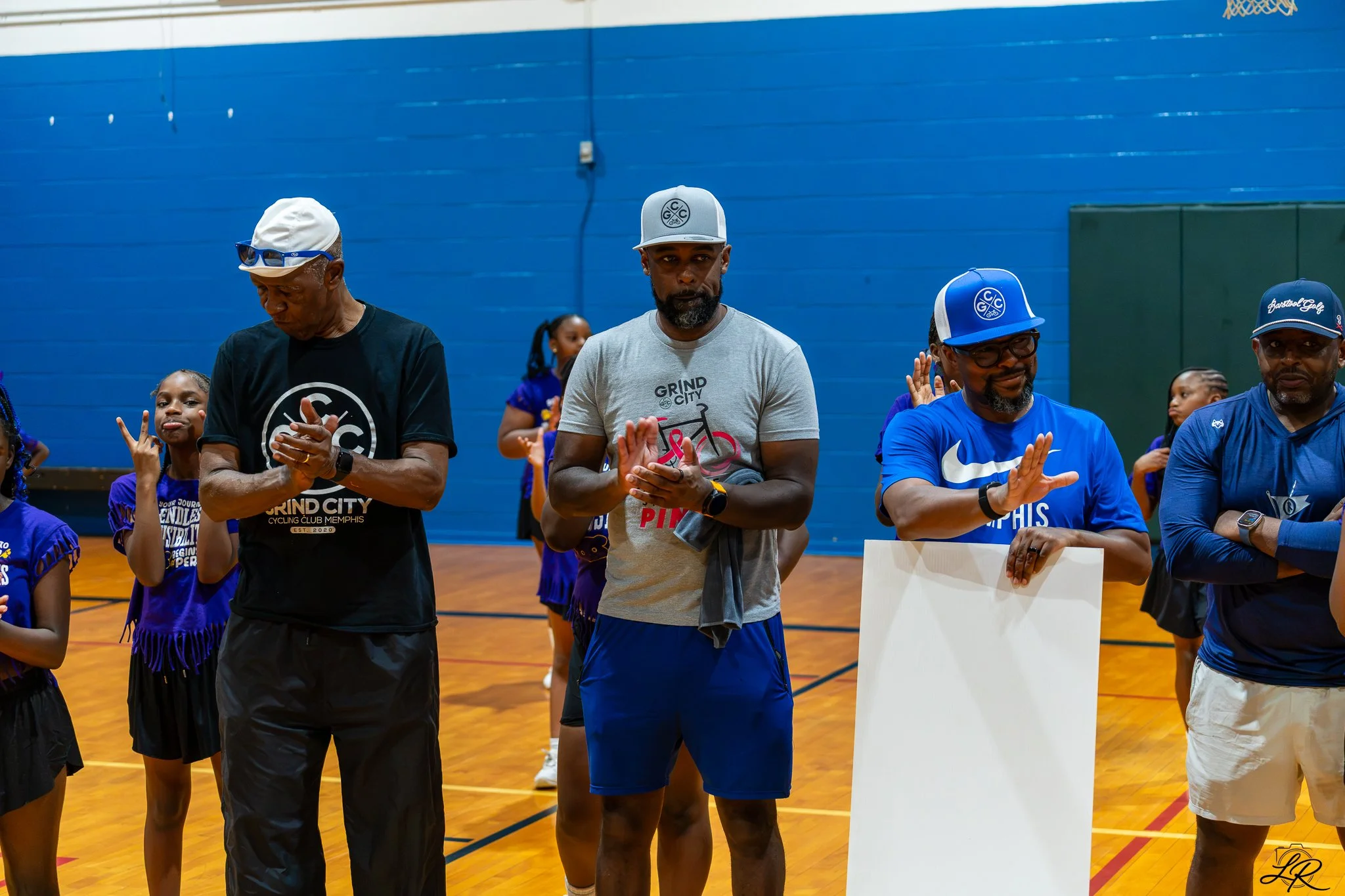 Group of men and young girls standing in a gymnasium with blue walls, some of the men wearing caps and t-shirts with print related to cycling and basketball.