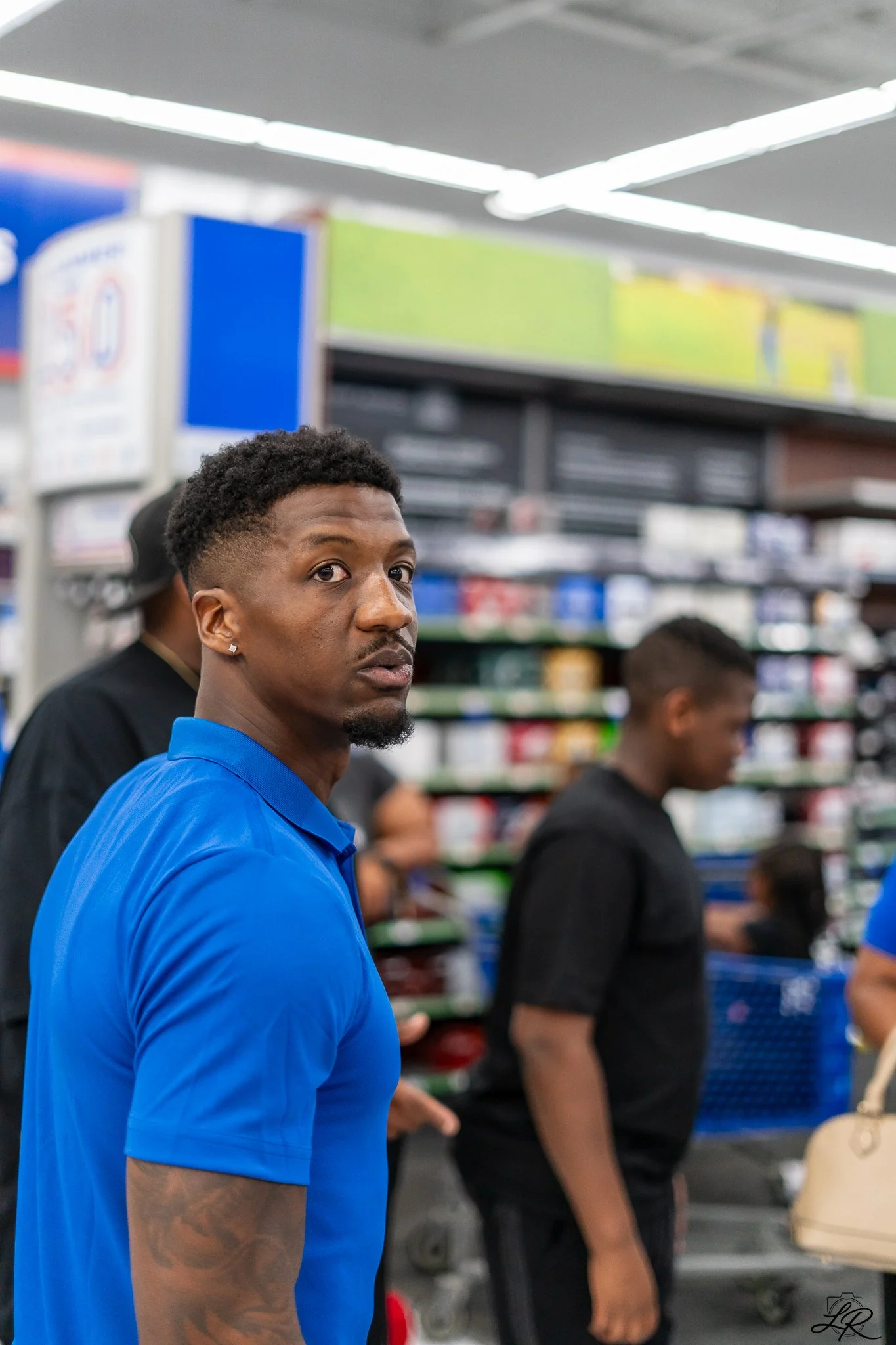A young man with curly hair, earrings, and a beard, wearing a blue shirt, looking towards the camera inside a store with shelves in the background.
