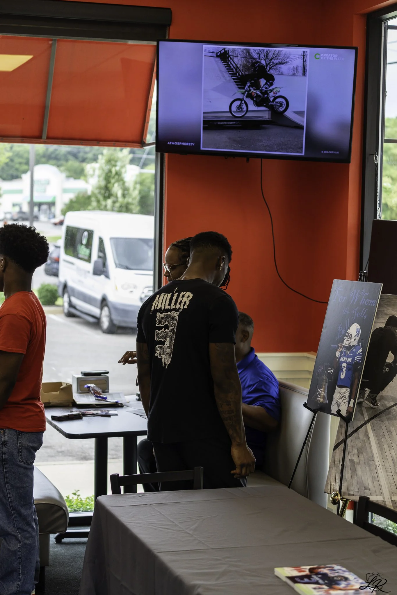 People gathered in a room with red walls and large windows, looking at a screen displaying a photo of a person riding a motorcycle down stairs outside. The room has tables, chairs, and posters on display.