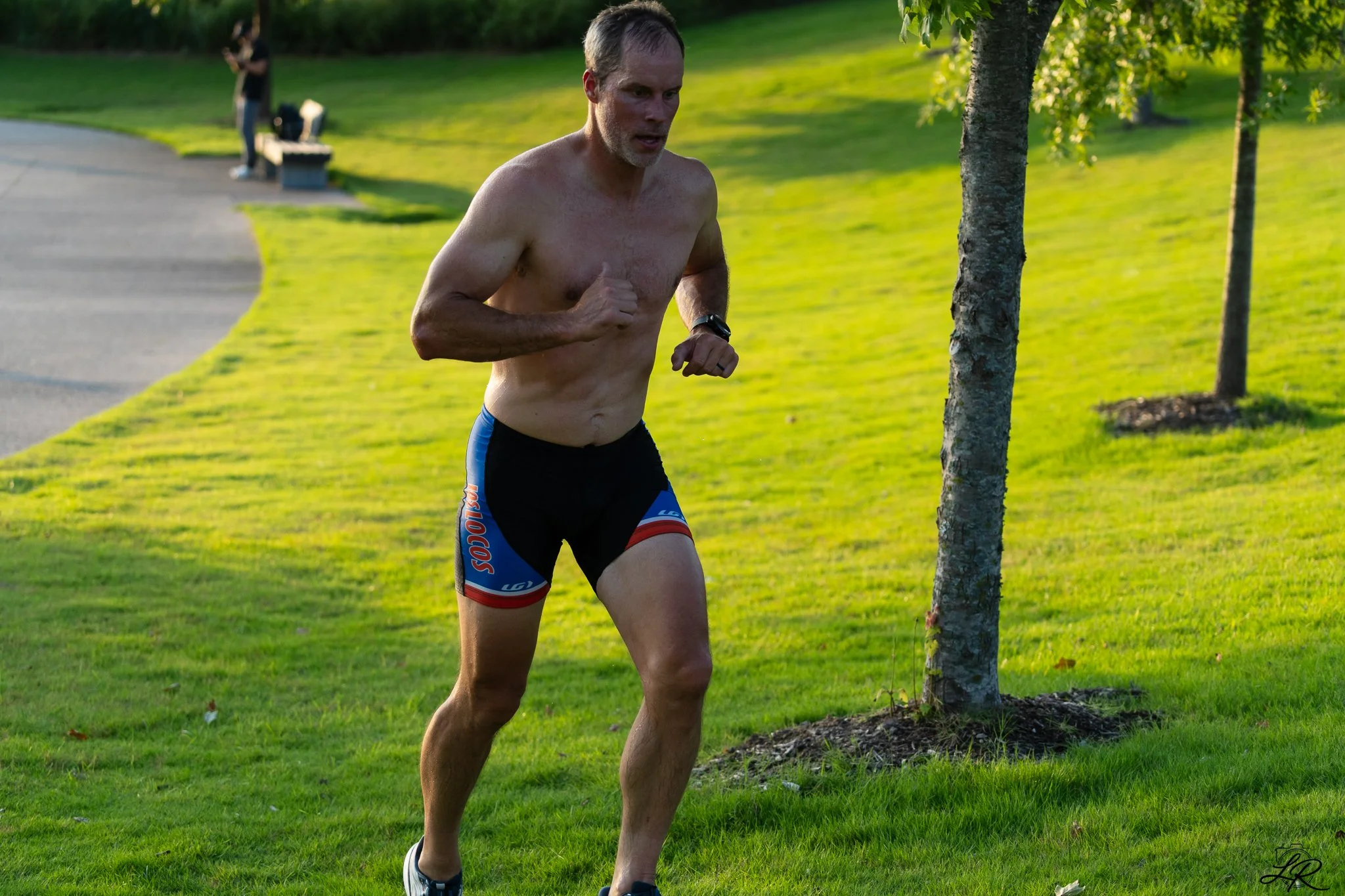 A shirtless man running outdoors on a grassy area near a tree, wearing black shorts with red, white, and blue accents.