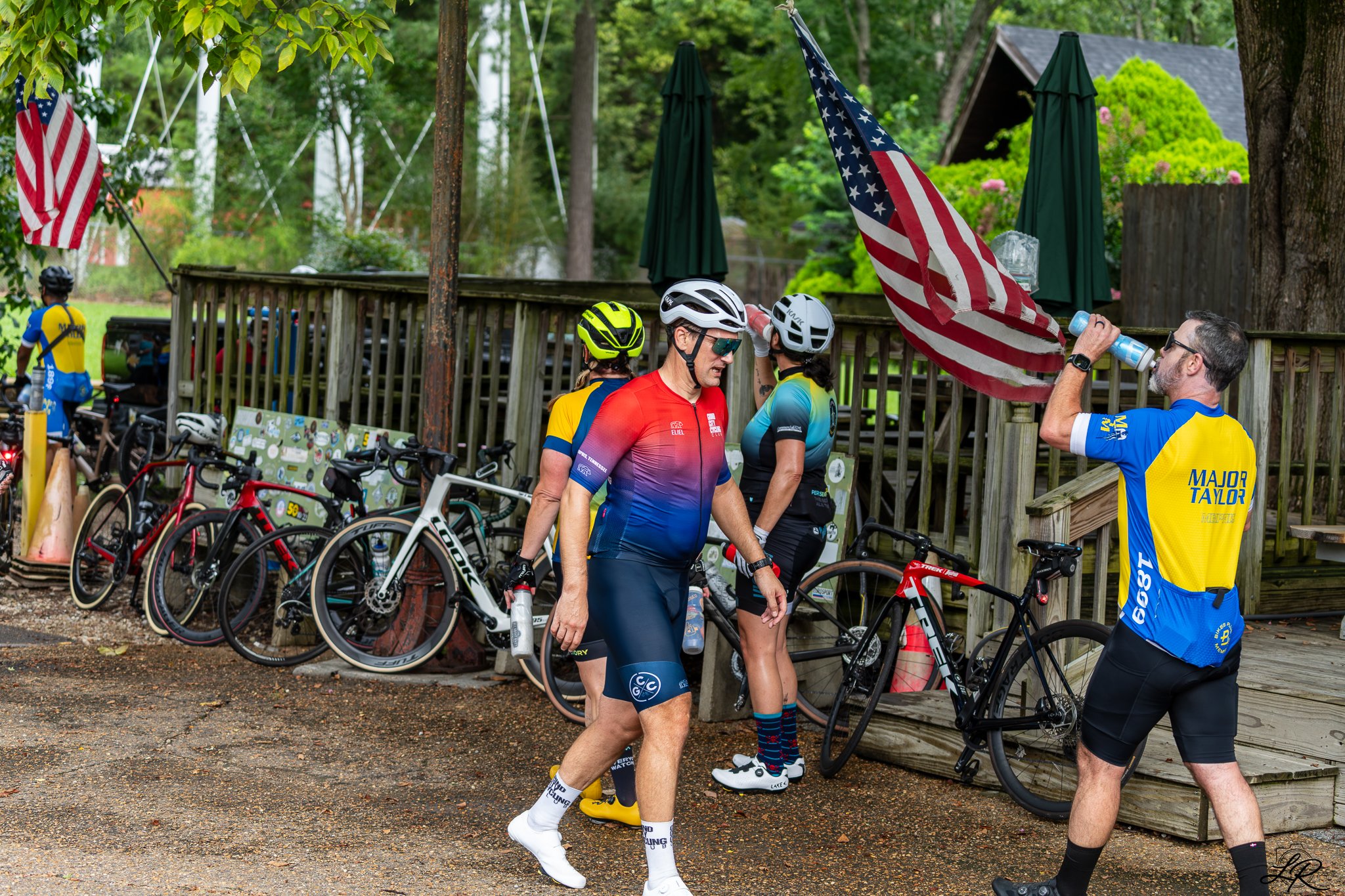 Cyclists gather near a wooden deck with American flags and green umbrellas, surrounded by trees, preparing for a bike race or event.