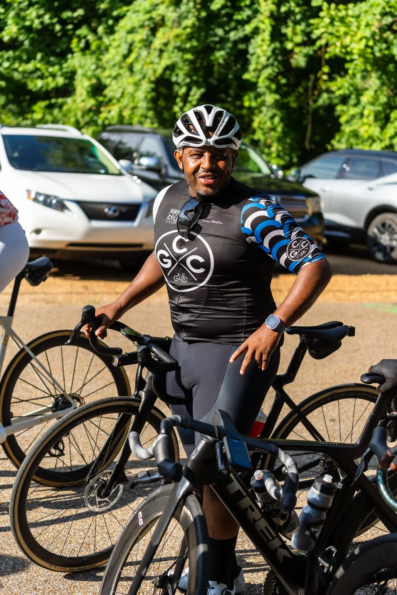 A man wearing a cycling helmet and black and blue cycling jersey standing with his bike outdoors, with several parked cars and green trees in the background.