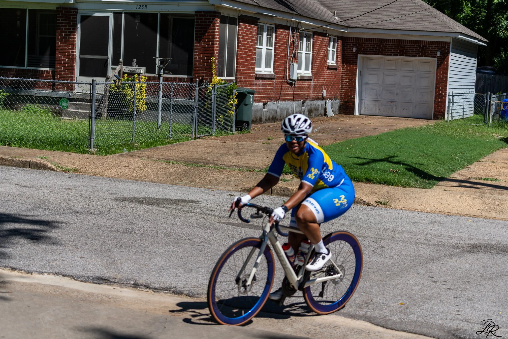 A young boy riding a bicycle on the street, wearing a white helmet and sunglasses, with a house and a lawn in the background.