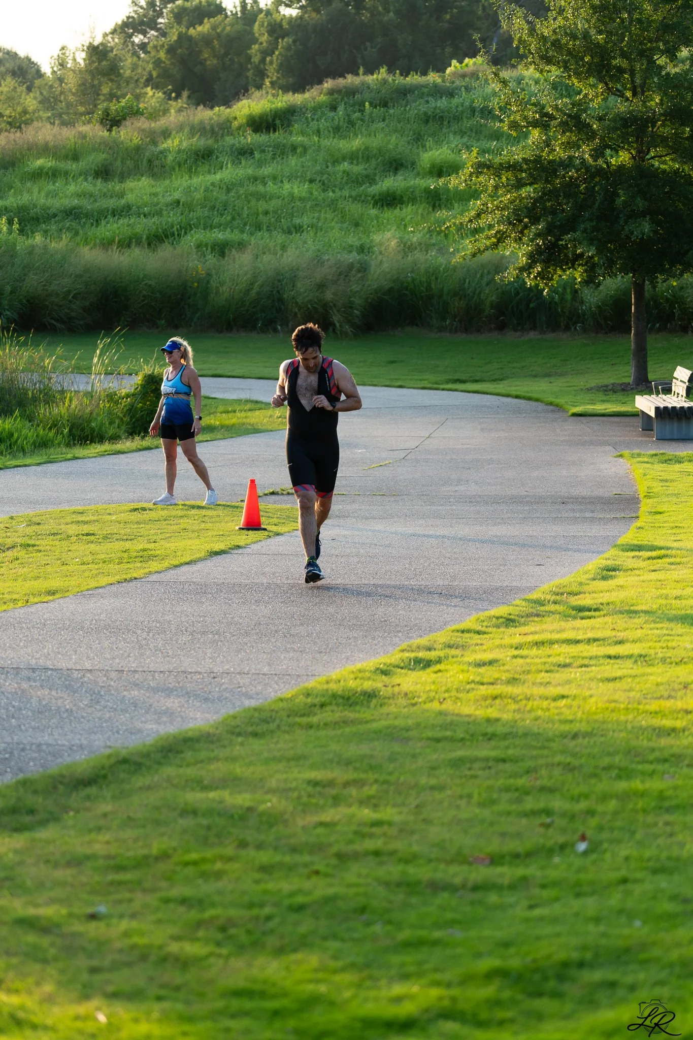 A man and woman jogging in a park with lush green grass, trees, and a walking path during the late afternoon or early evening.