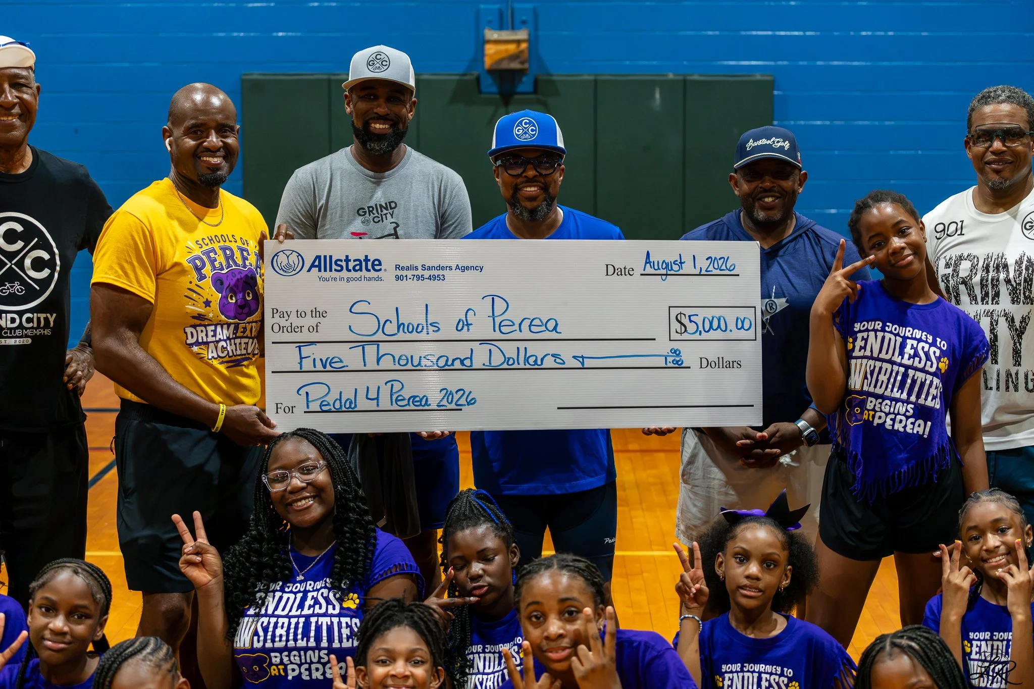 Group of smiling African American adults and children in a gymnasium holding a giant check for $5,000 made out to Schools of Perea, dated August 1, 2026.