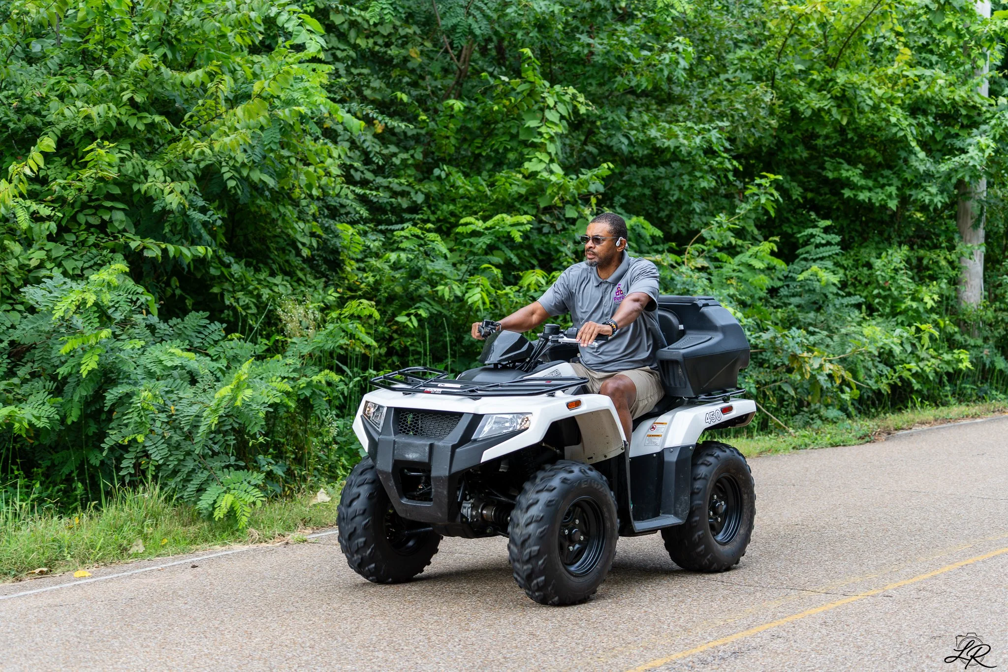 A man riding an all-terrain vehicle (ATV) on a paved path beside a dense green forest, wearing sunglasses and a gray polo shirt.