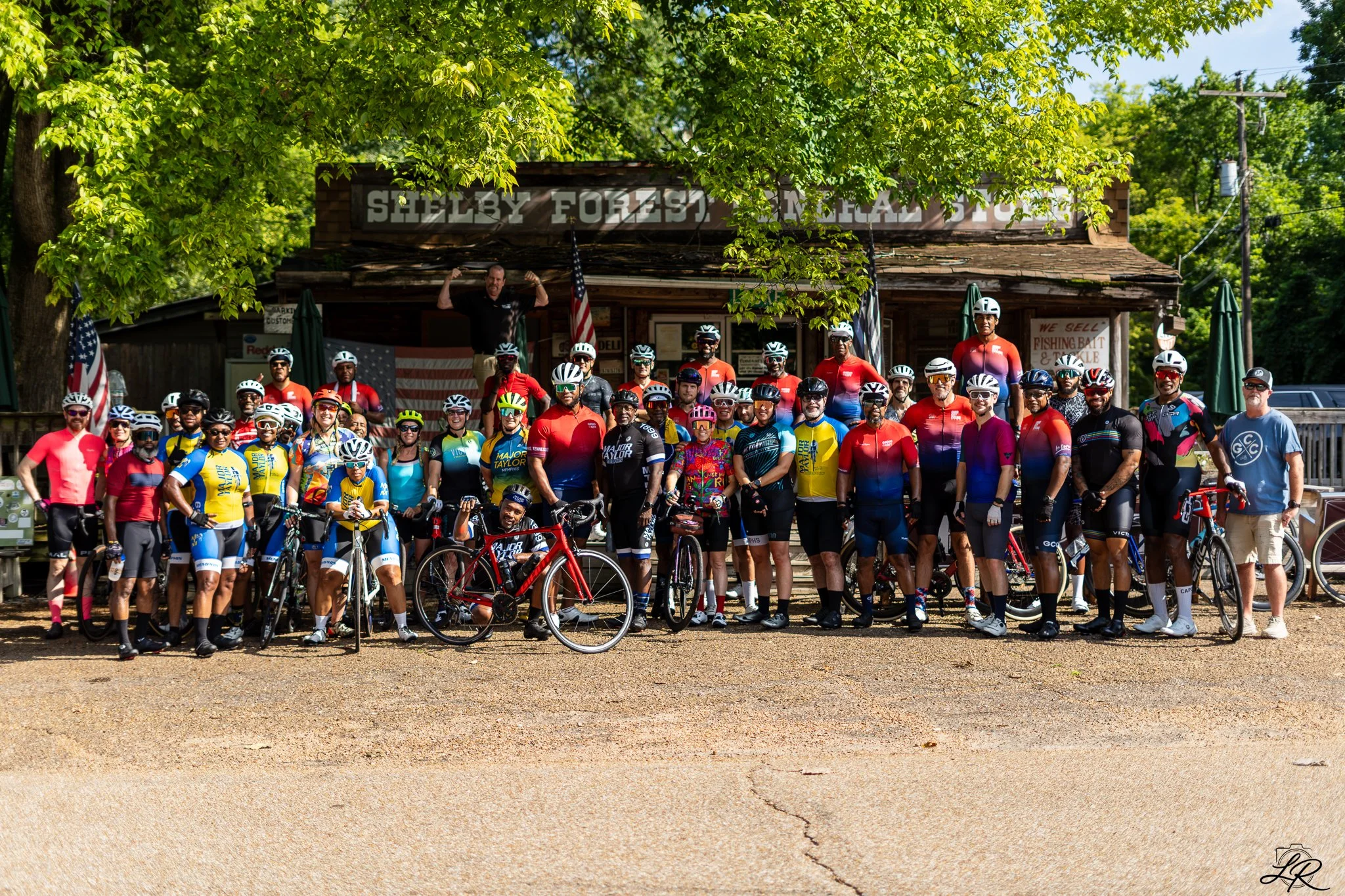 Group of cyclists posing in front of a rustic wooden building with a sign that reads Shelby Forest General Store. The cyclists are wearing colorful jerseys and helmets, and some are holding their bikes. There is a man standing on the porch of the sto