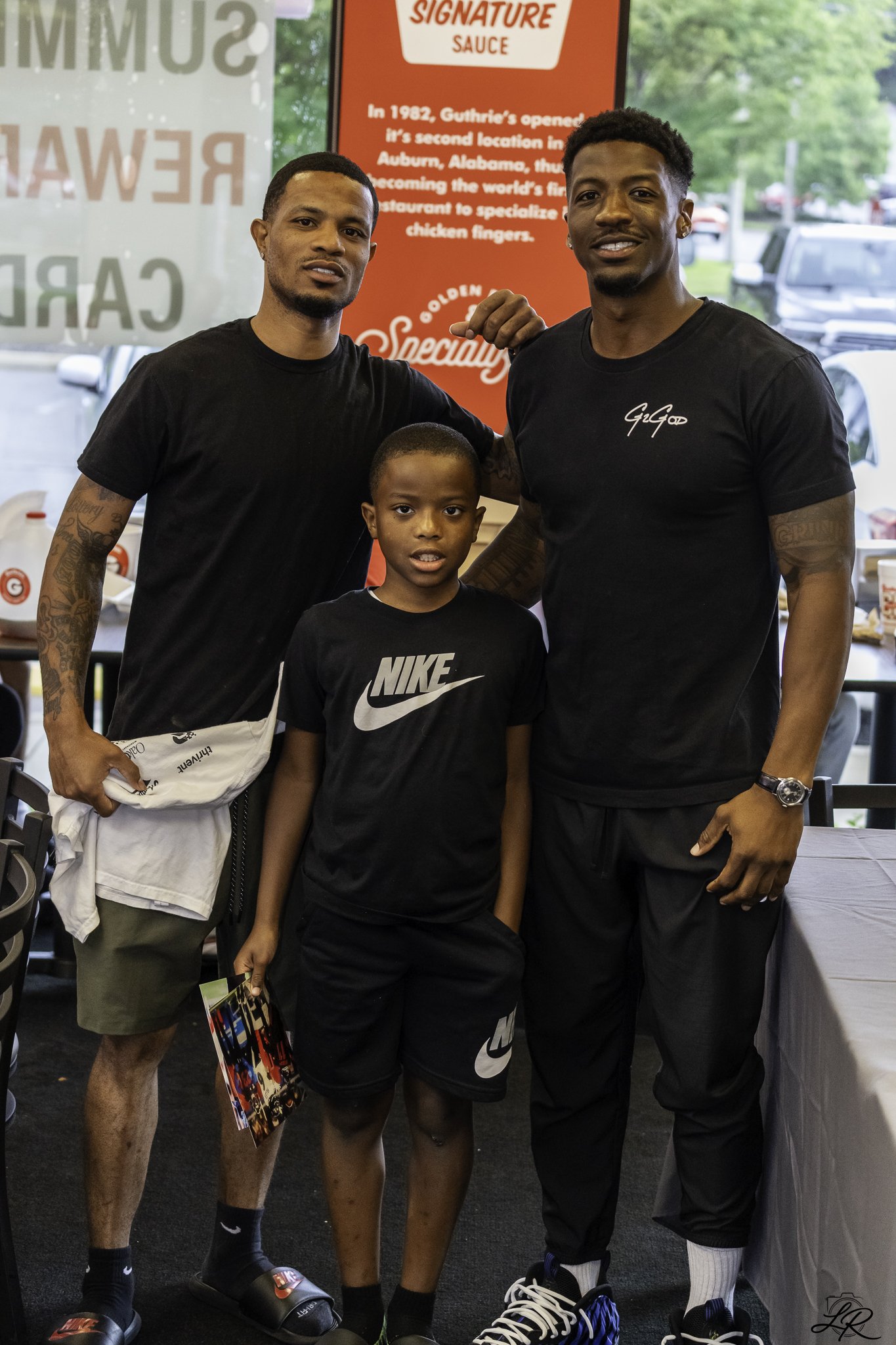 Three males and one young boy standing inside a restaurant, posing for the camera. Behind them is a red sign with white text about the restaurant's history and signature sauce. The man on the right is flexing his arm and wearing a black Guga t-shirt,