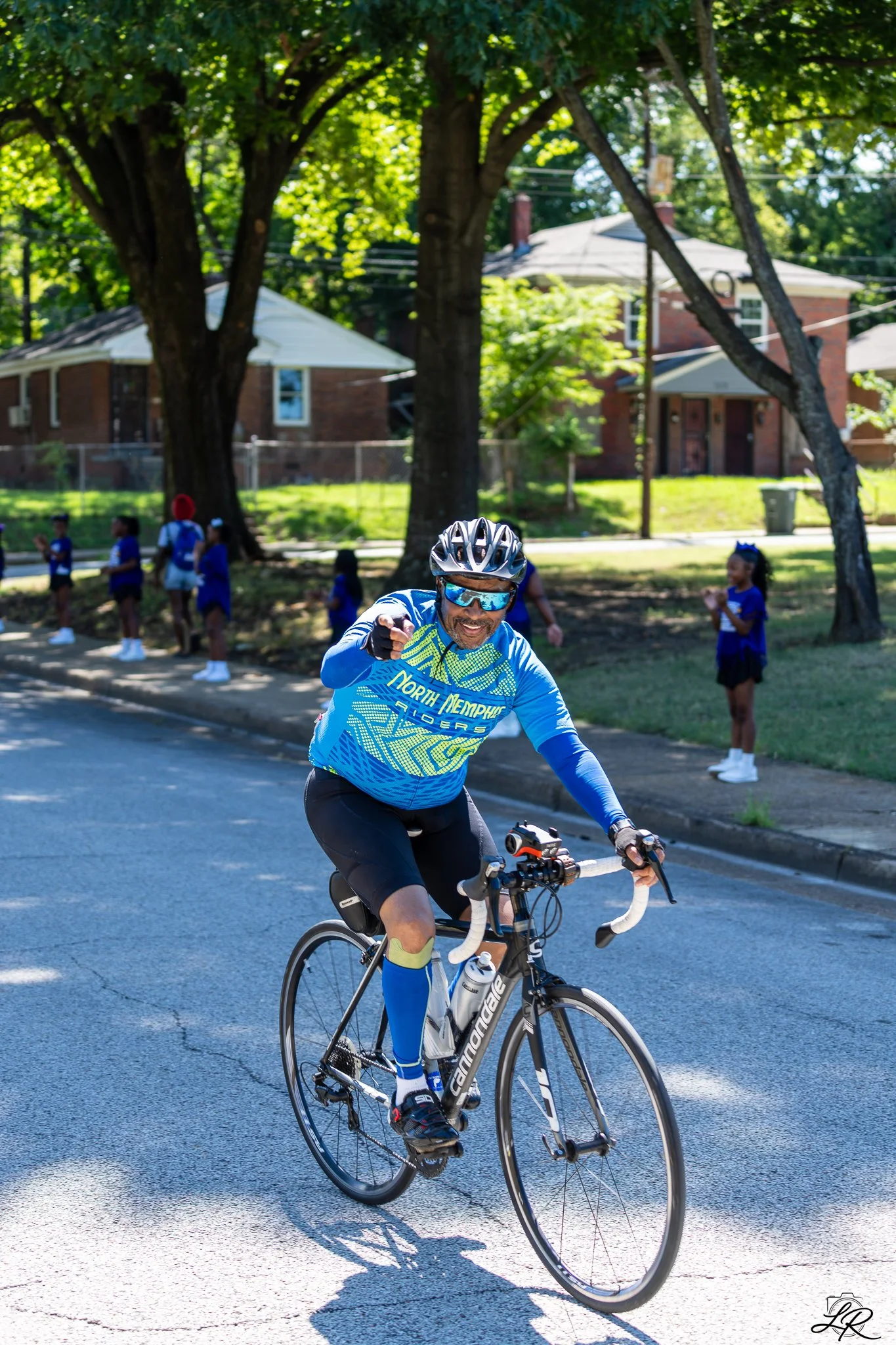 A man riding a bicycle on a street in a neighborhood with houses and trees in the background. The man is wearing a blue cycling jersey, black shorts, sunglasses, a helmet, and gloves. Several children in blue uniforms are standing on the sidewalk beh