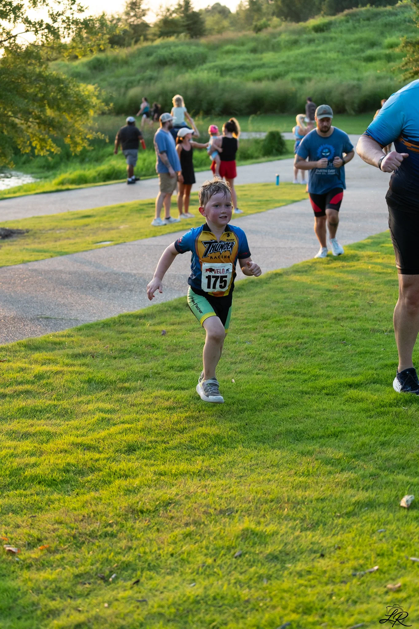 A young boy running outdoors in a park or nature trail during daytime. He is wearing a racing bib with the number 175, a blue and yellow racing jersey, and light-colored shorts. There are people in the background walking and standing near a grassy ar