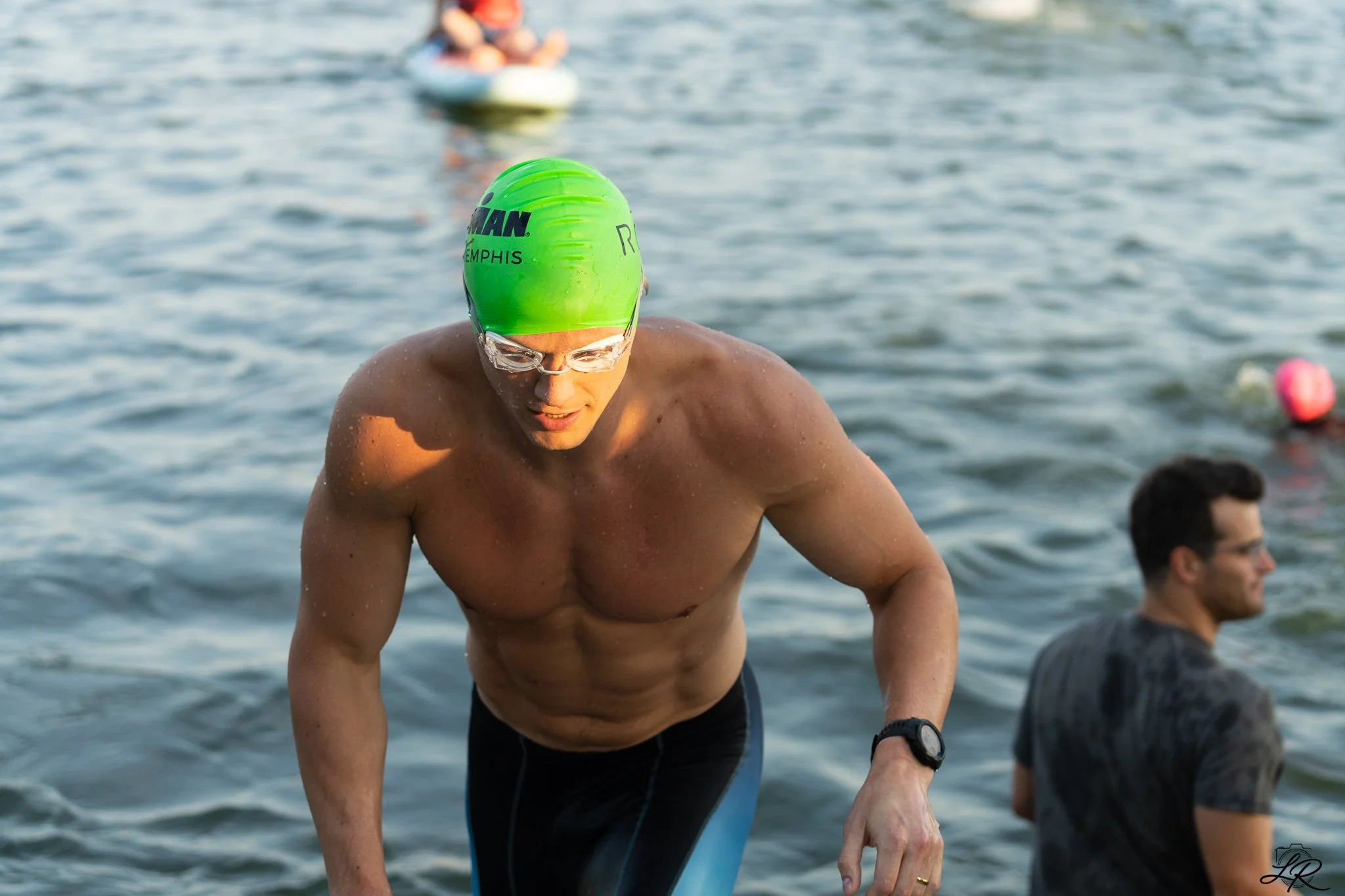 A male swimmer with a green swim cap and goggles emerging from the water, with other swimmers and buoys in the background.
