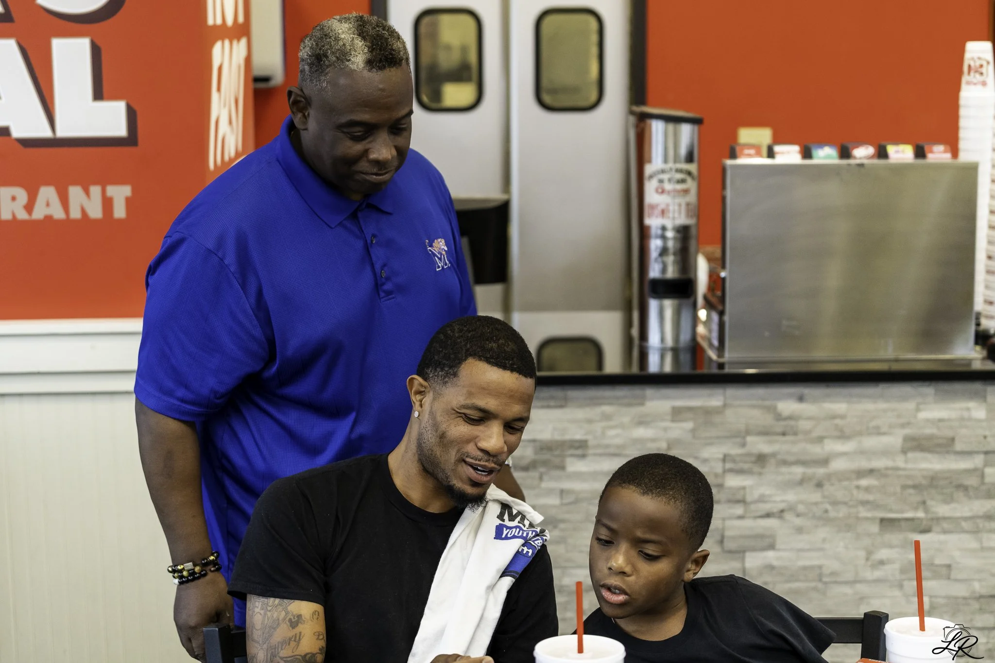 People gathered around a table in a fast-food restaurant. A man with gray hair and a blue shirt stands behind a young man with tattoos and a black shirt, both looking at something on the table. The background features red walls, drink machines, and a
